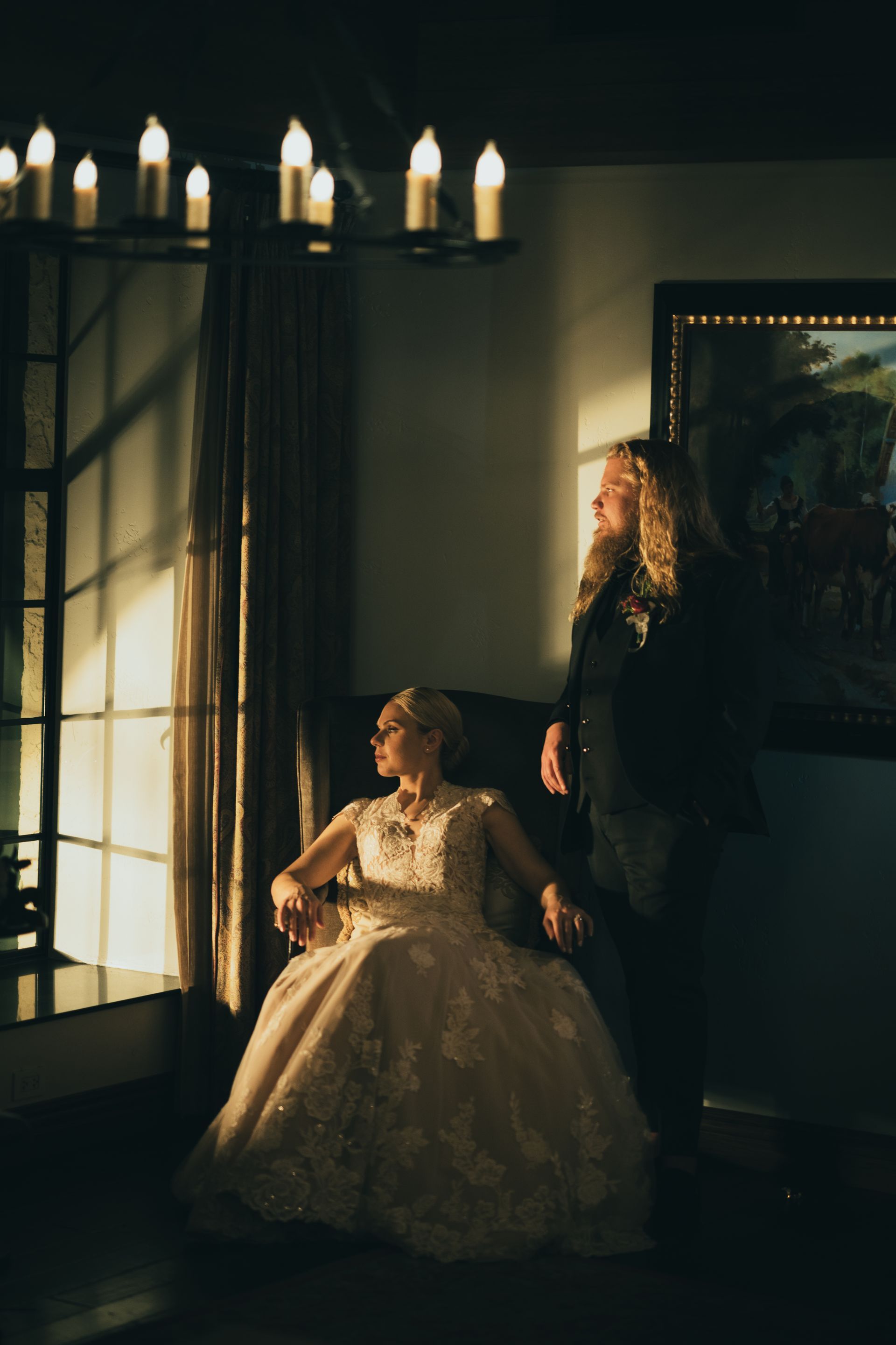 Woman in gown seated by window, man beside her; chandelier, painting, and room with golden light.