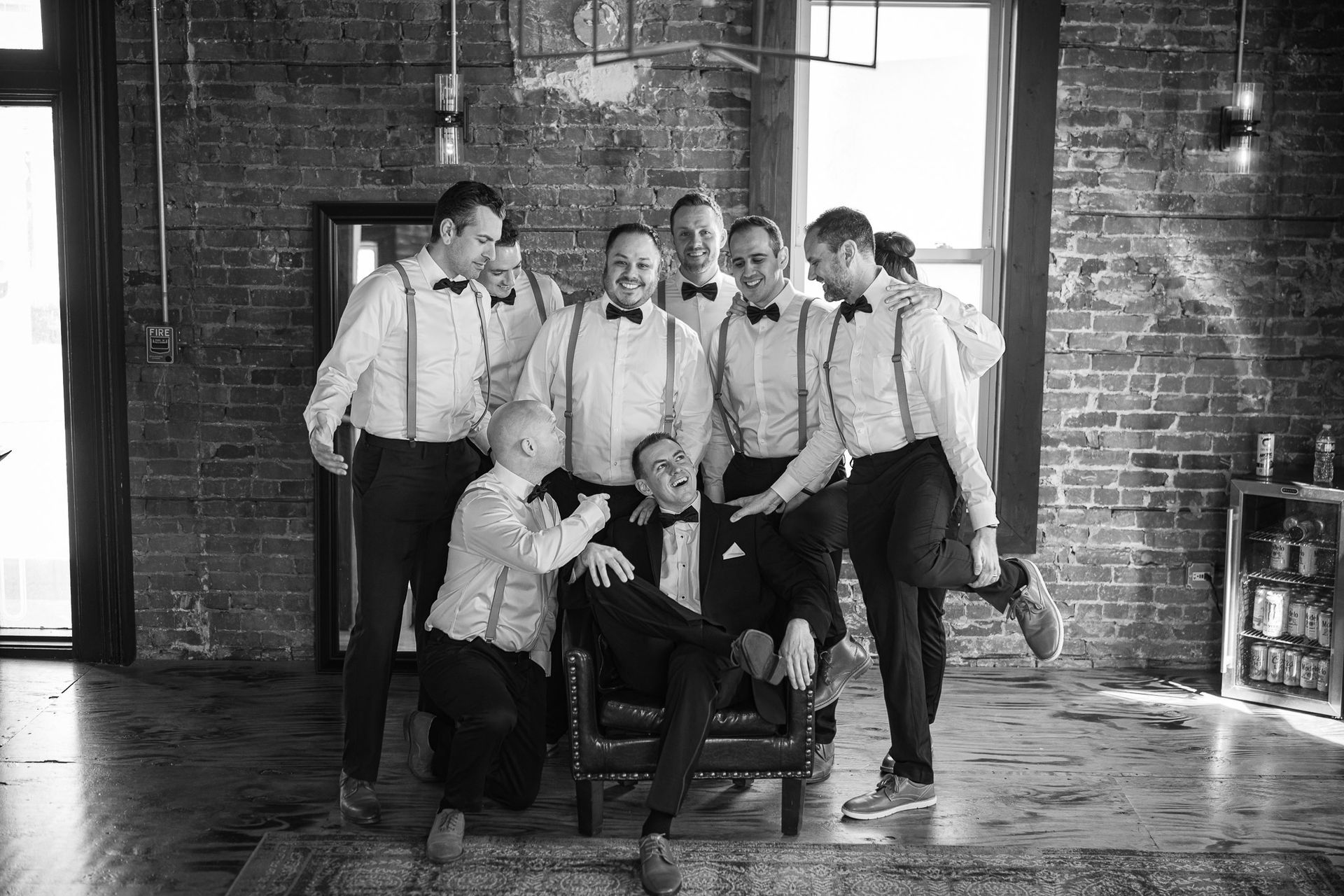 Groomsmen in suspenders surround a man in a tuxedo, posing for a photo in a brick-walled room.