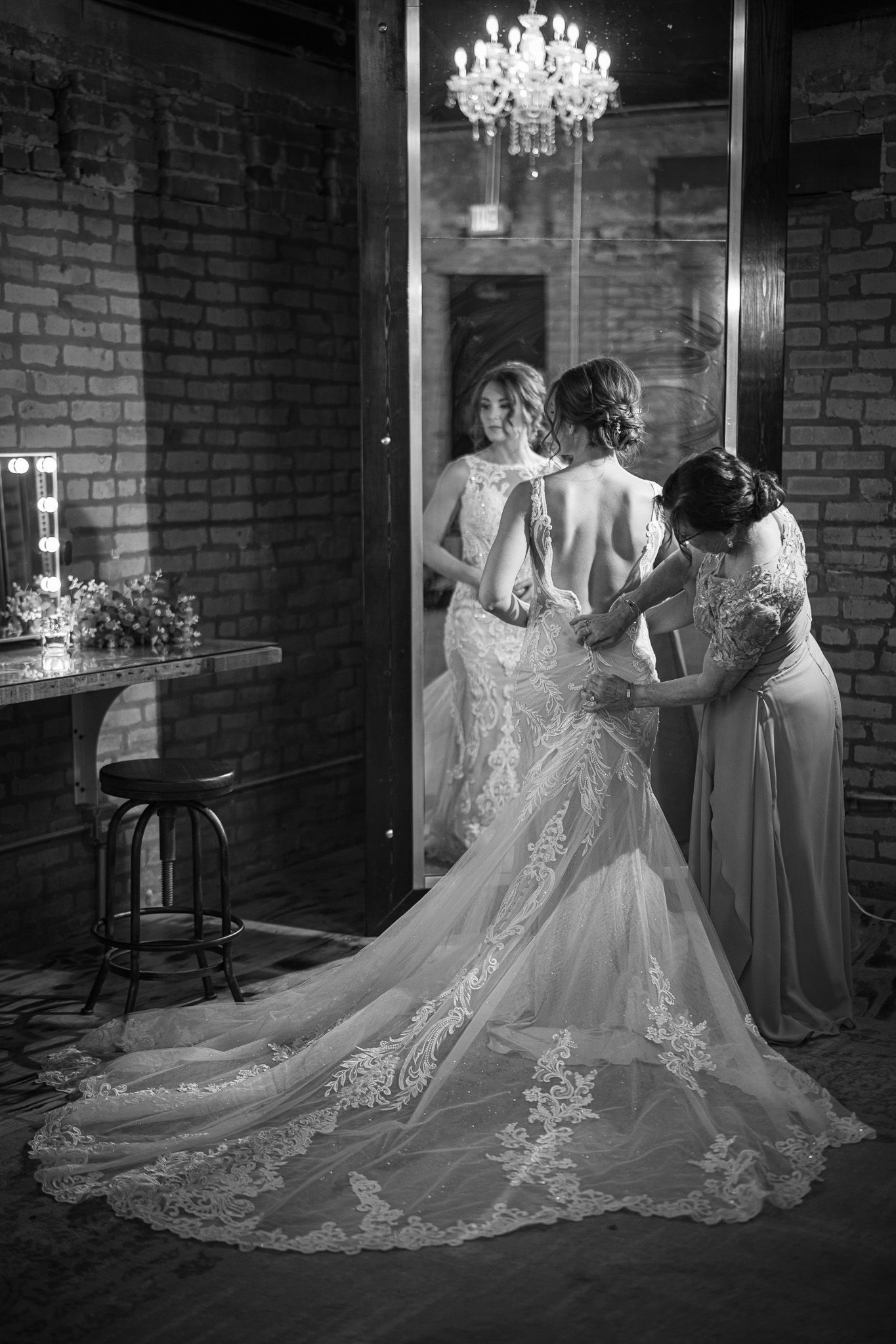 Bride in wedding dress, being helped by two women in a room with a large mirror and brick walls.