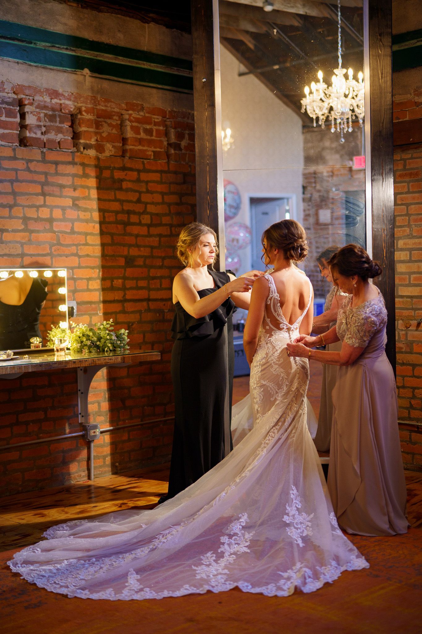 Bride in wedding dress, being helped by two women near a large mirror in a brick-walled room.