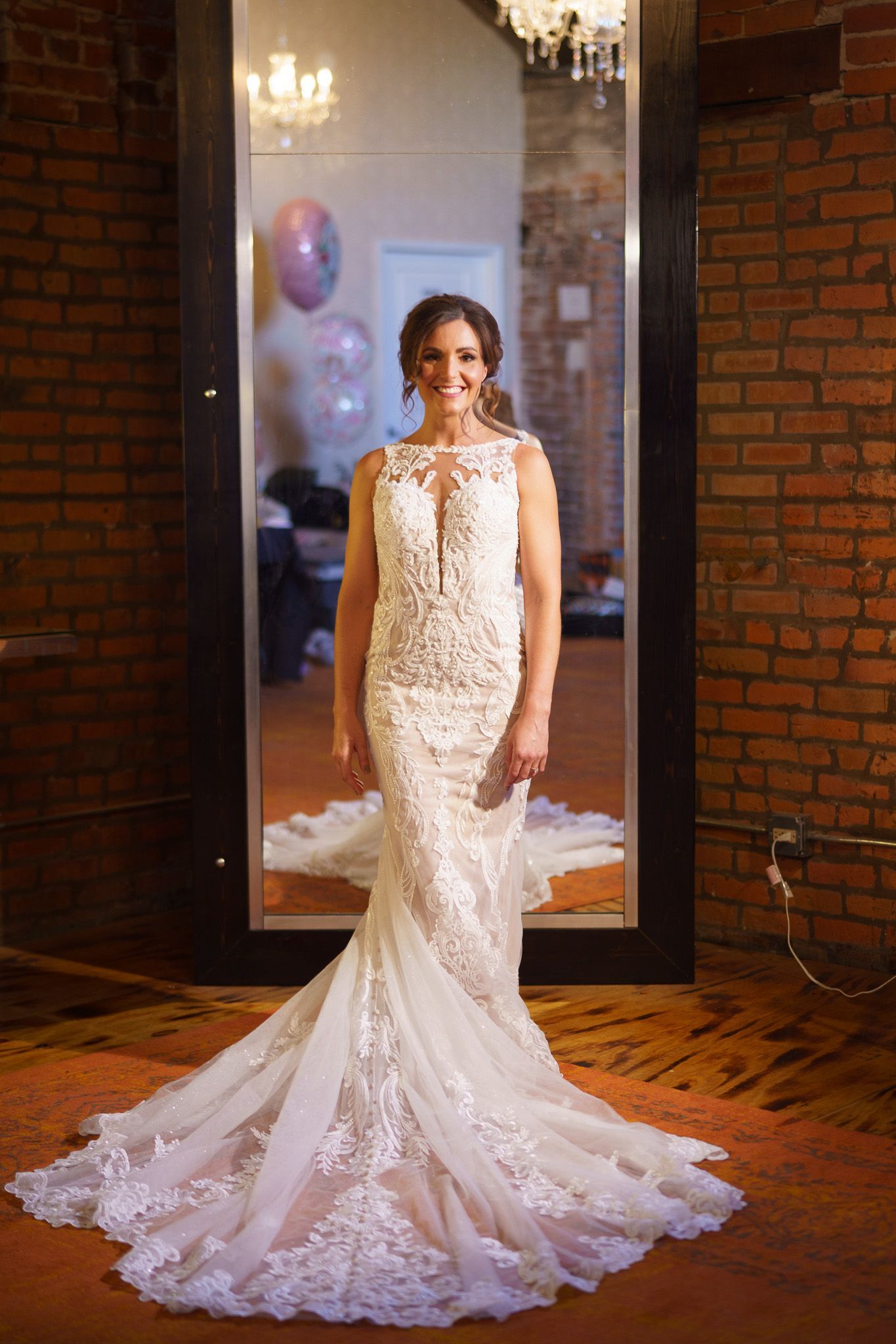 Woman in a white lace wedding dress poses in front of a mirror, brick wall backdrop.