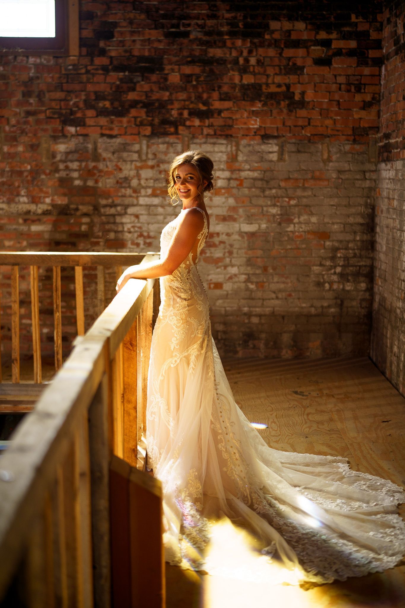 Woman in wedding dress leans on wooden railing, sunlight on brick wall.