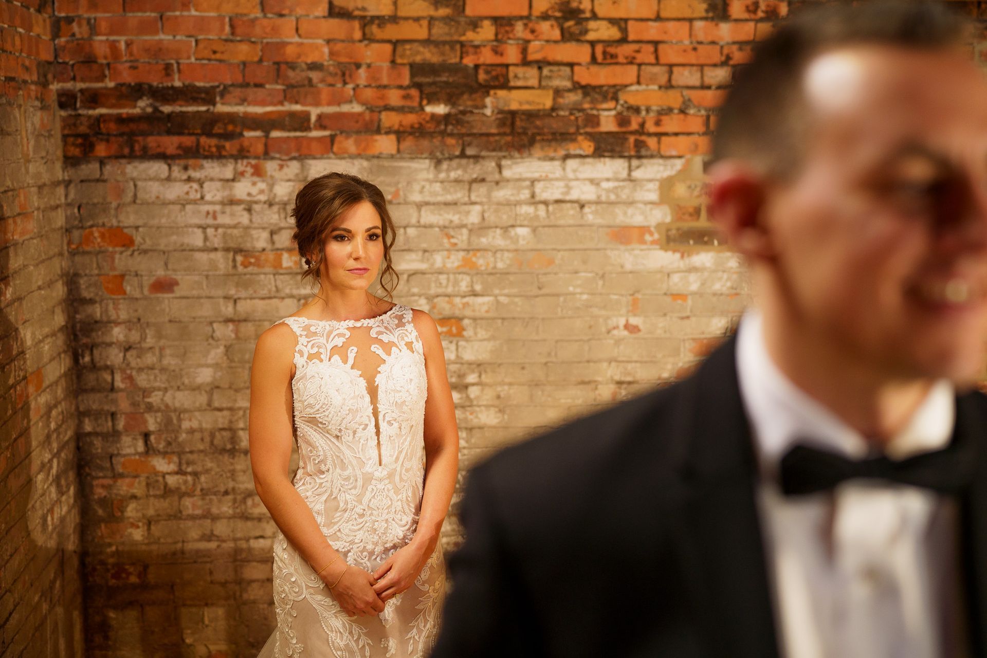 Bride in white lace gown stands before a brick wall as groom is slightly out of focus in the foreground.