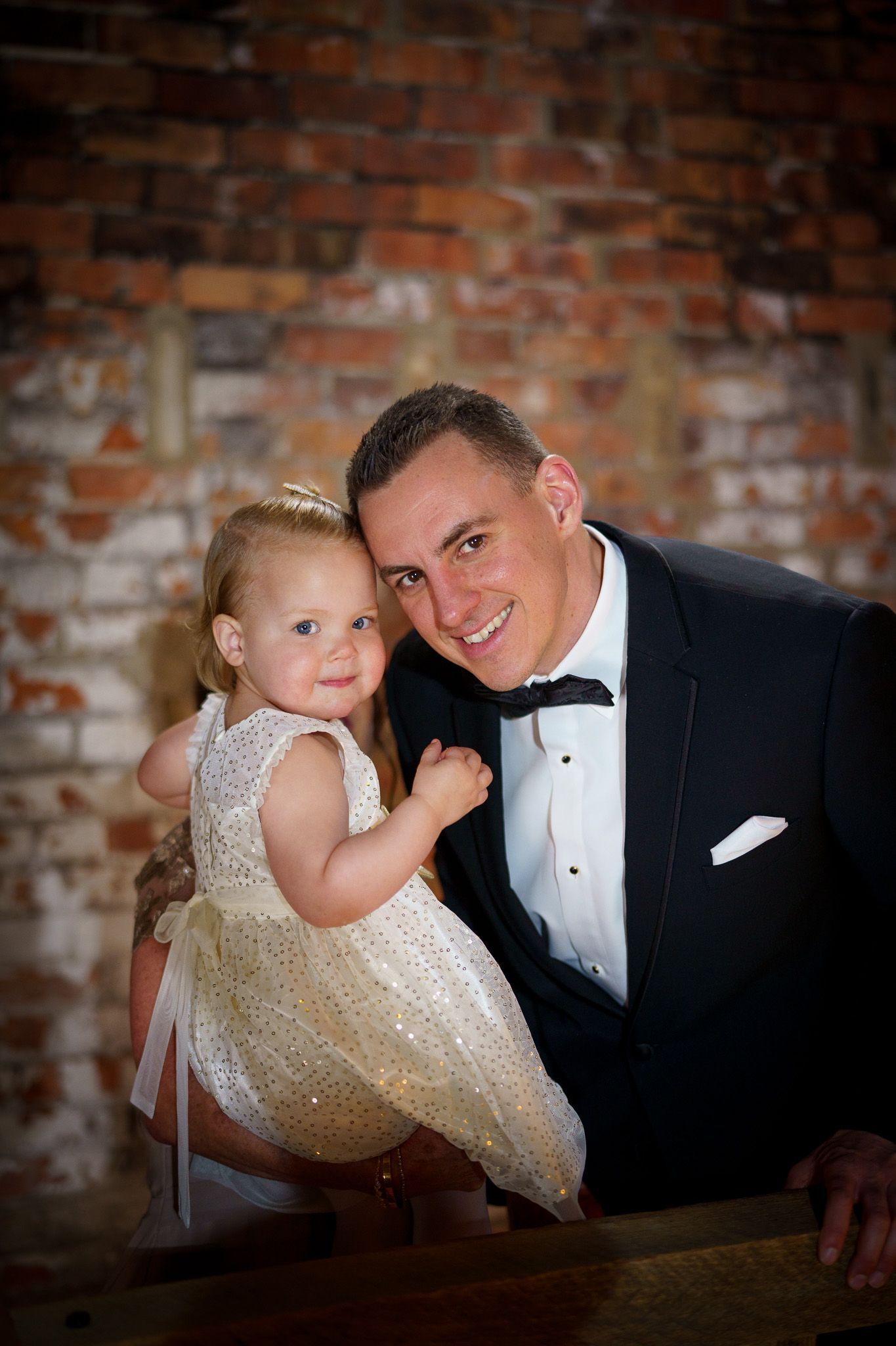 Man in tuxedo holding a toddler in a gold dress; brick wall background.