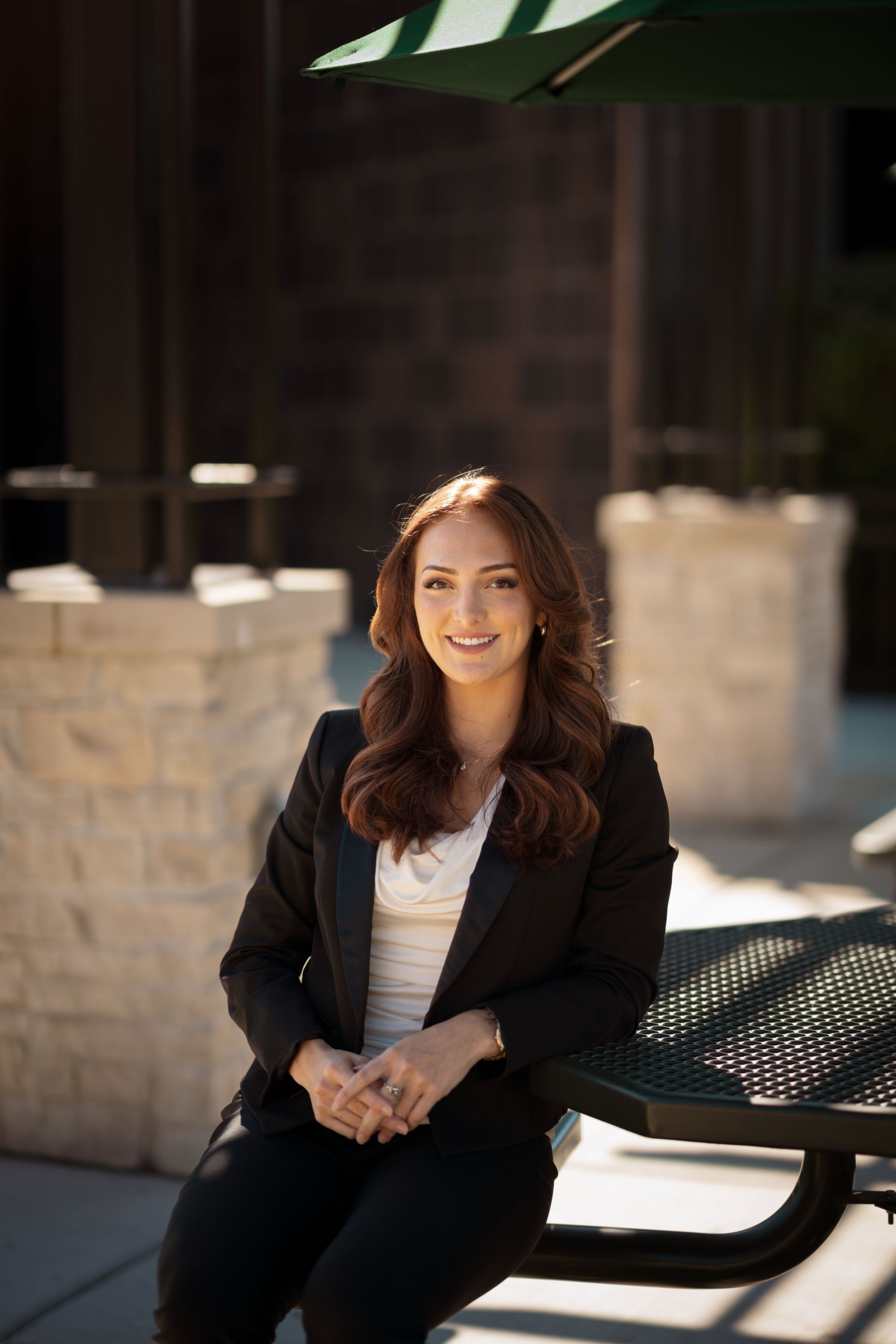 Woman in a black blazer sits at an outdoor table, smiling.