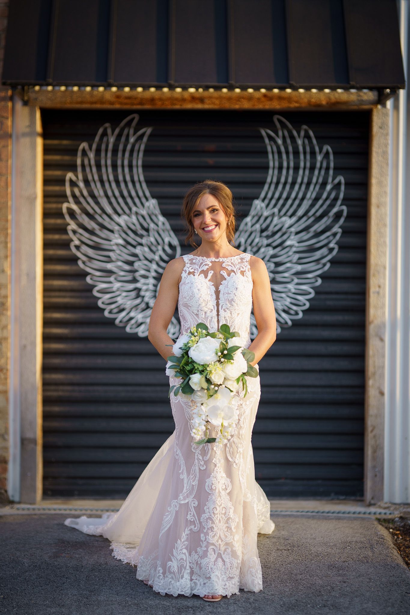 Bride in a lace gown holding flowers, posing in front of an angel wing mural on a dark metal door.