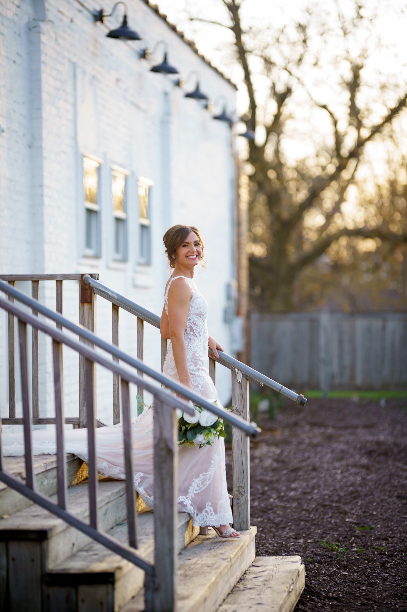 Bride in white wedding dress smiles while descending wooden stairs of a white building, holding flowers.
