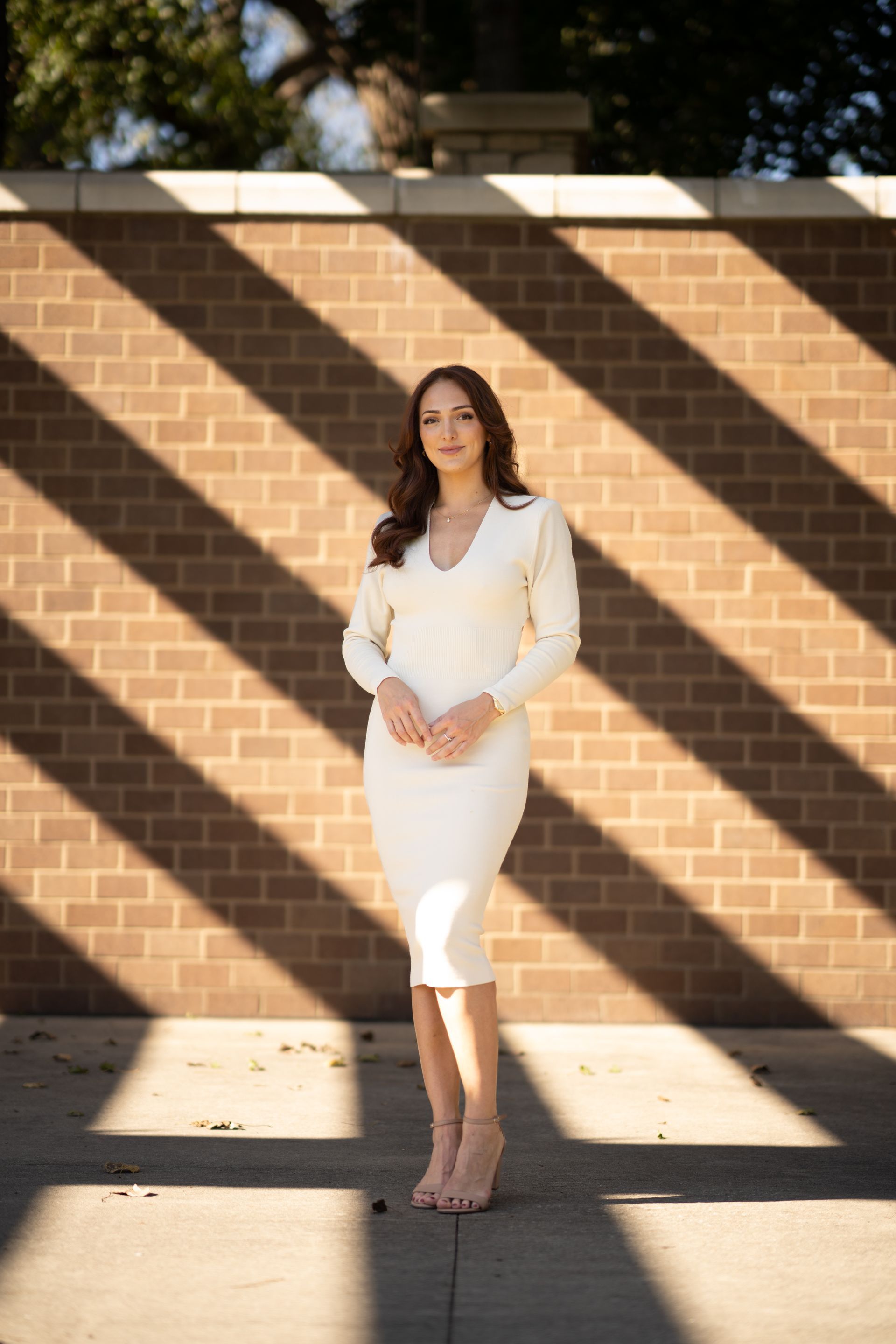 Woman in a white dress and heels poses against a brick wall with shadow stripes.