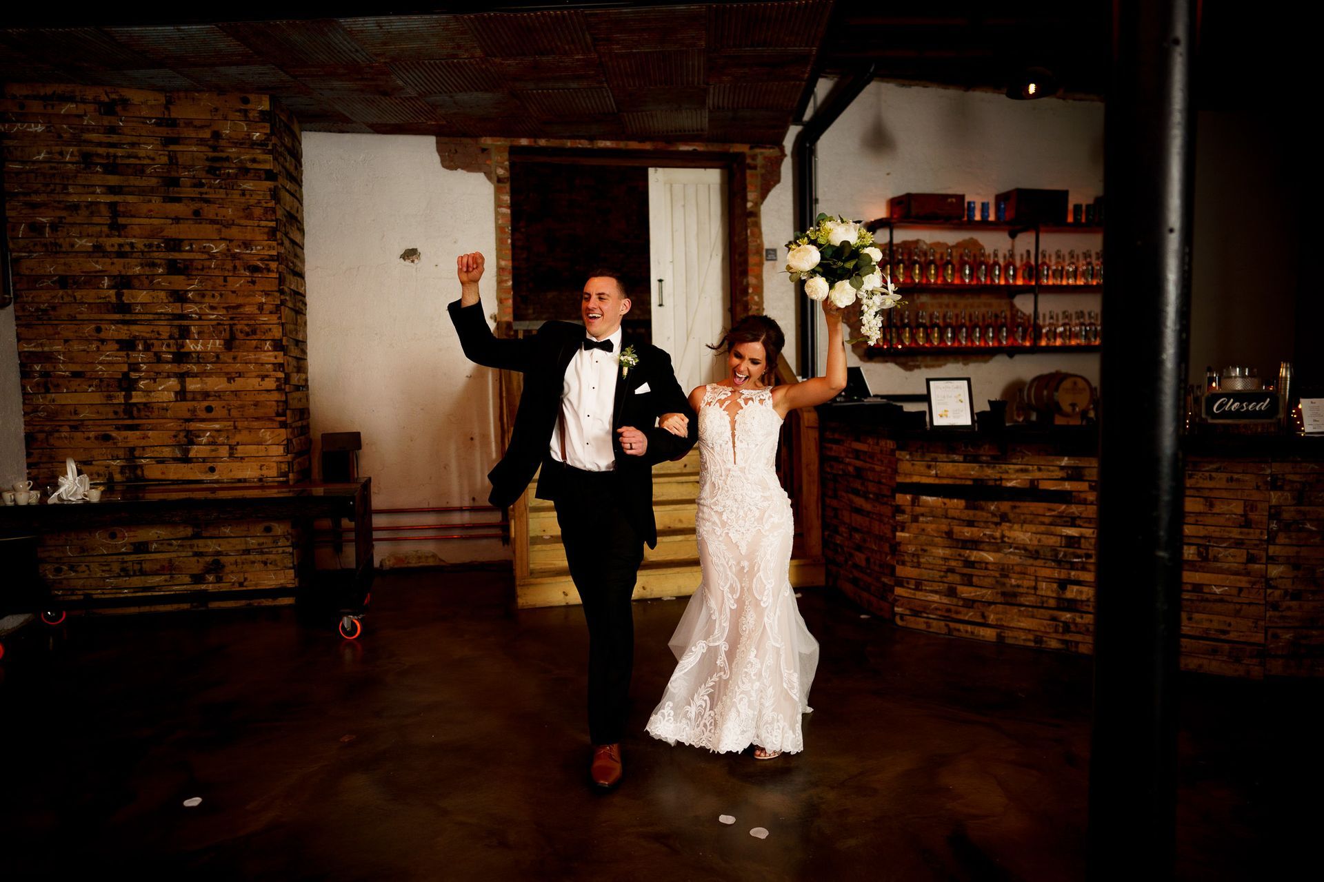 Newlyweds celebrate, raising arms. Woman in lace dress holds bouquet; man in tuxedo. Rustic brick interior.