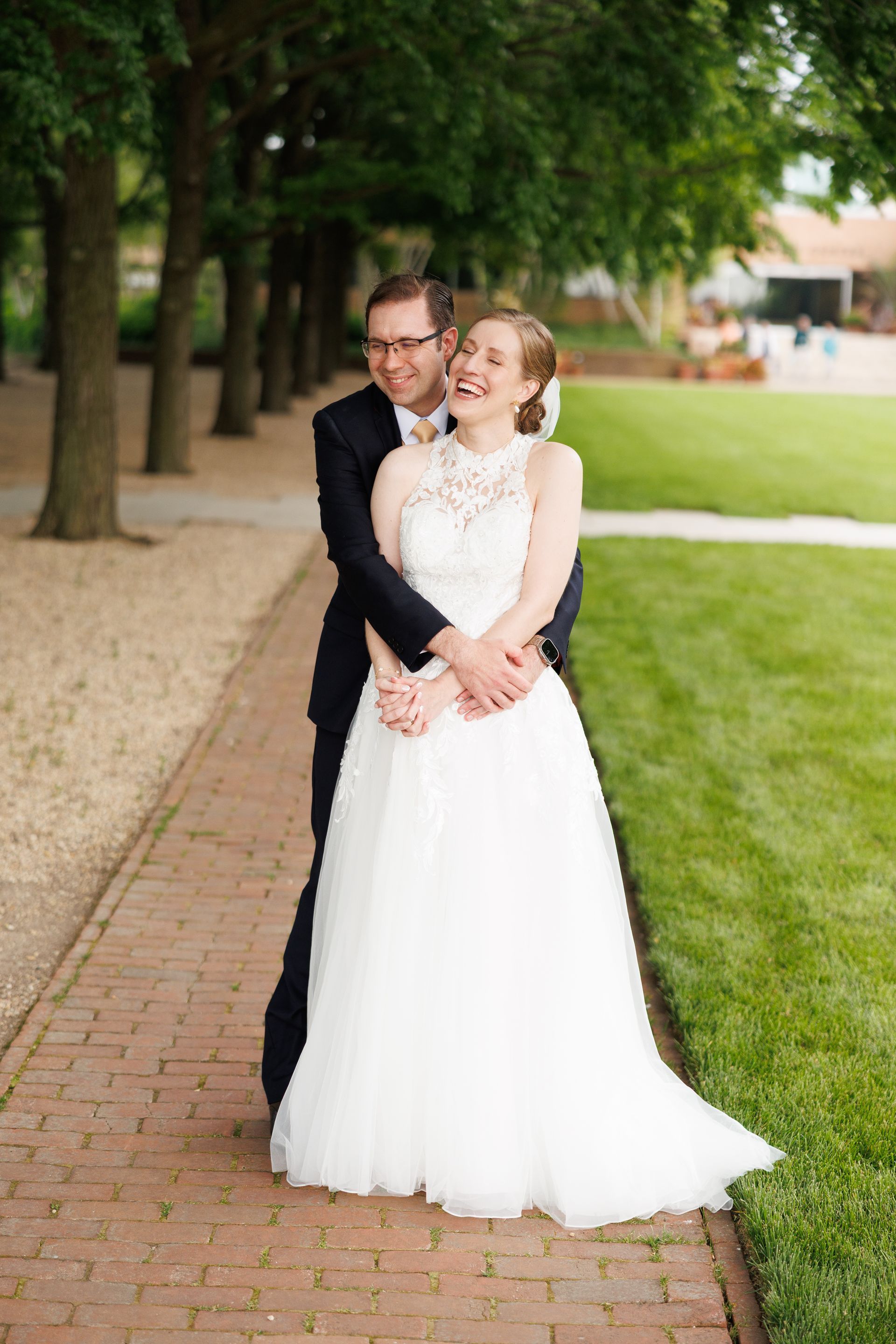 Bride and groom embrace on a brick path, she in white dress, he in black suit, smiling, green lawn backdrop.