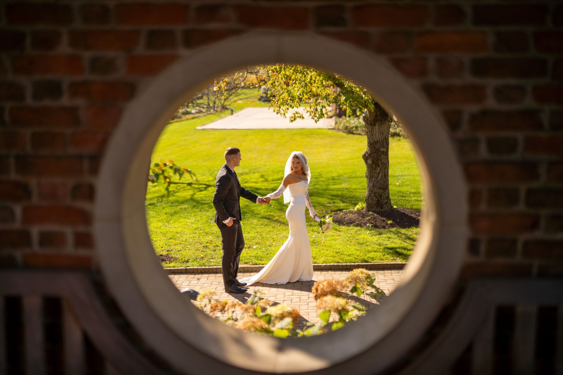 Couple holding hands, framed by a circular opening in a brick wall, overlooking a sunny green lawn.