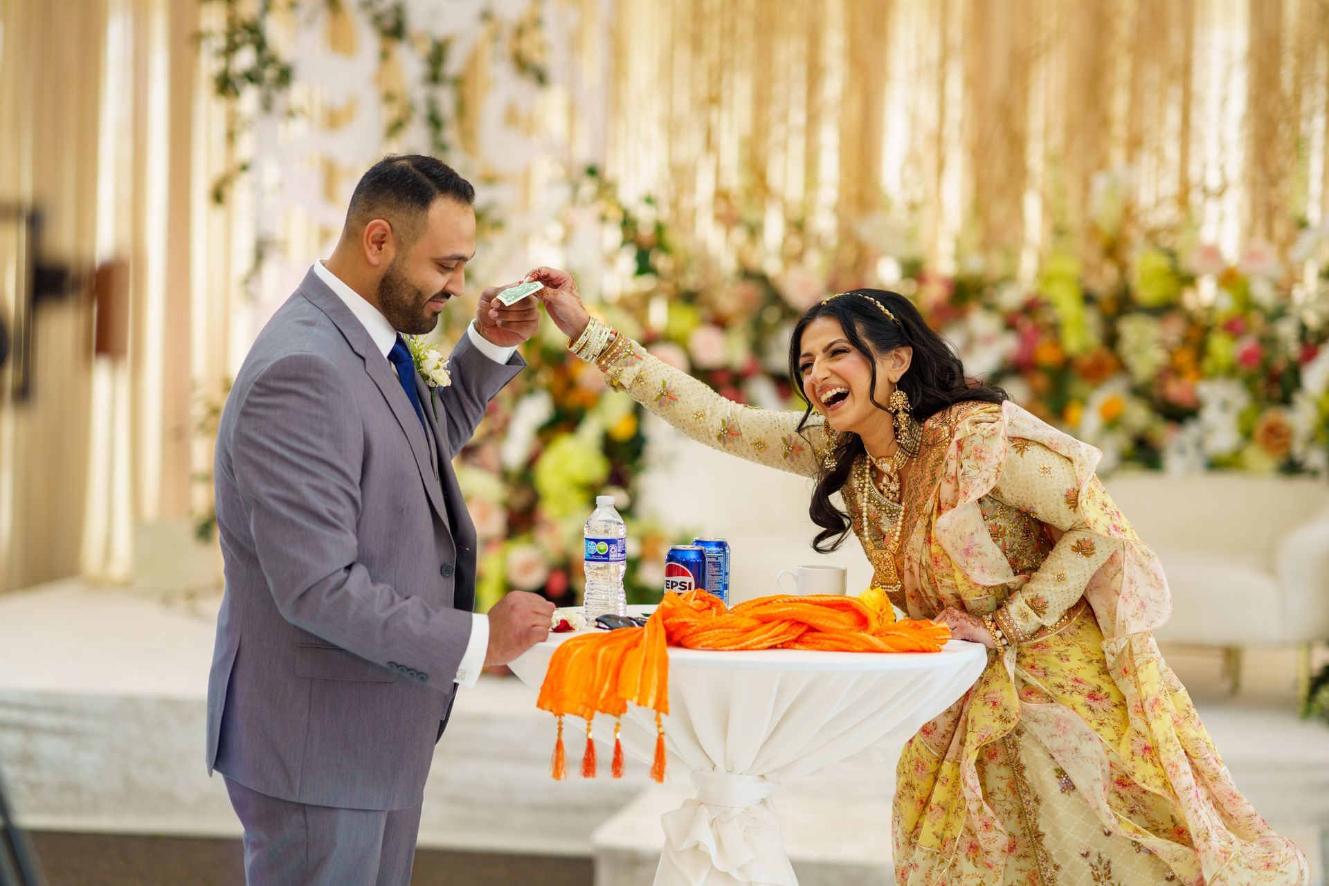 Man and woman at event; she playfully reaches for something. He wears a gray suit, she a yellow and gold dress, surrounded by flowers.
