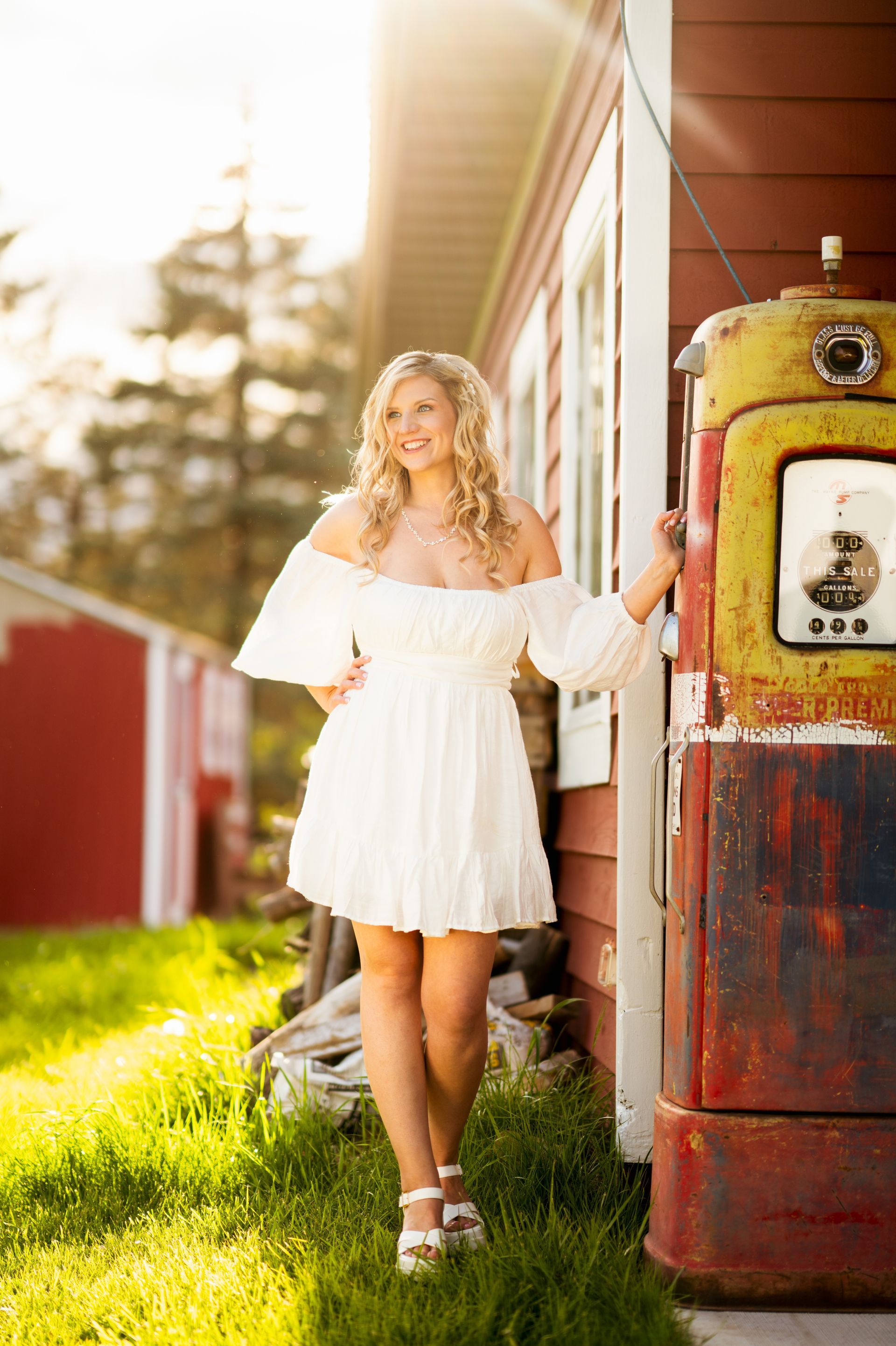 Woman in white dress leans on vintage gas pump by red building, sunlit outdoor scene.