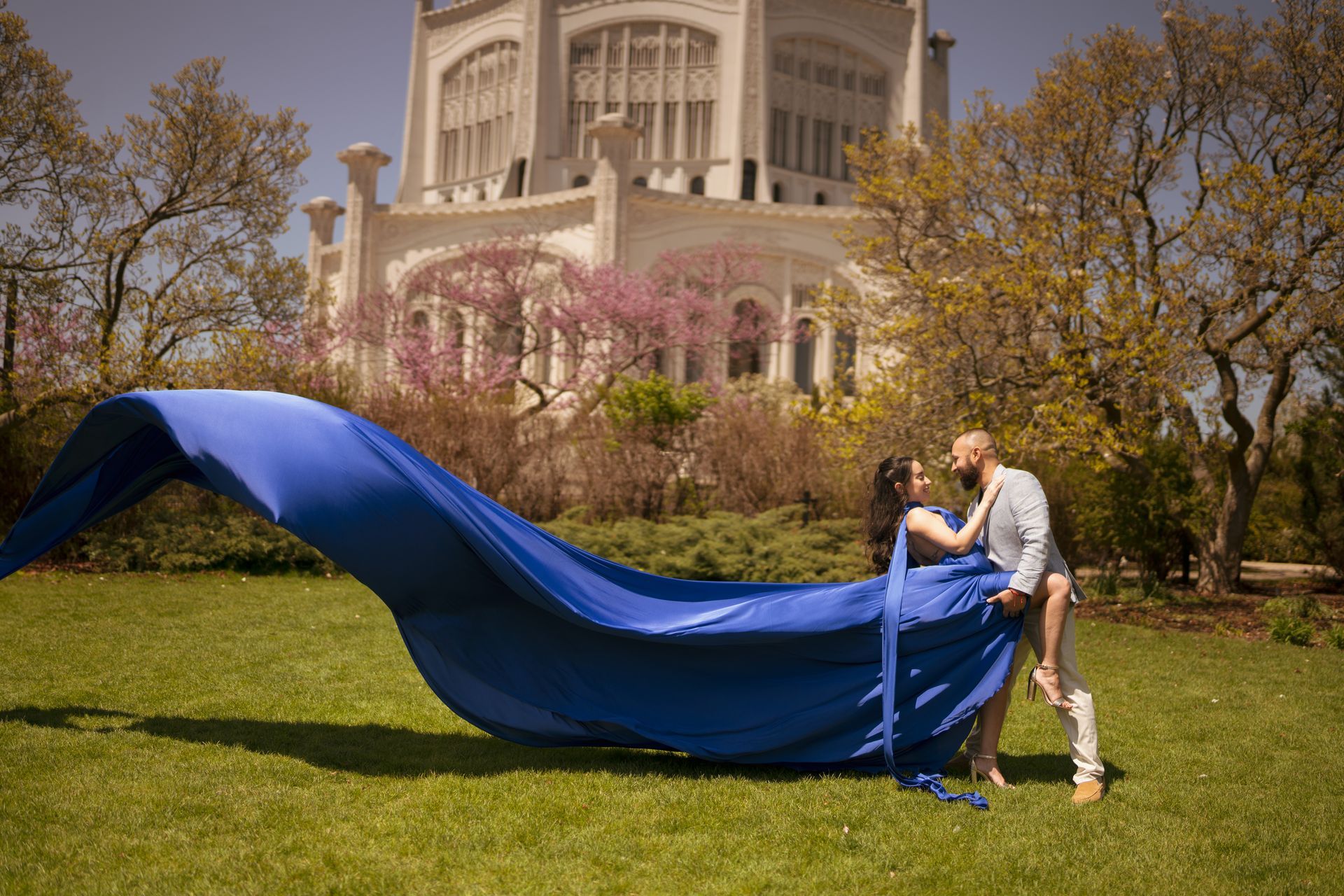 Couple in embrace with blue fabric, in front of a white building with trees and green grass.