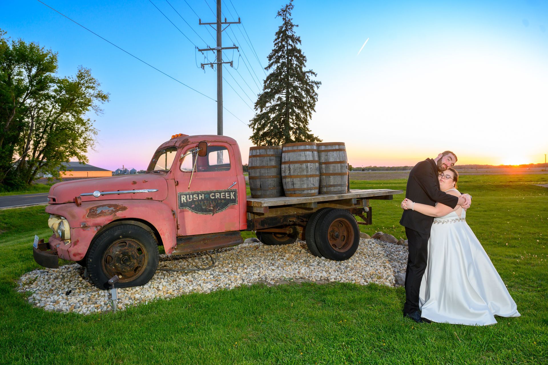 Couple embracing beside an old red truck with barrels, sunset background.