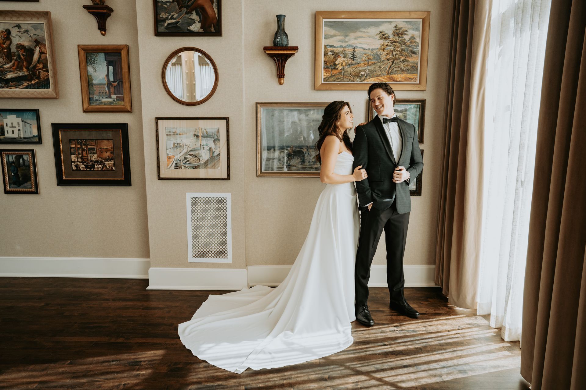 Bride and groom pose near a window. A wall of framed art decorates the wall. The bride wears a long white gown.