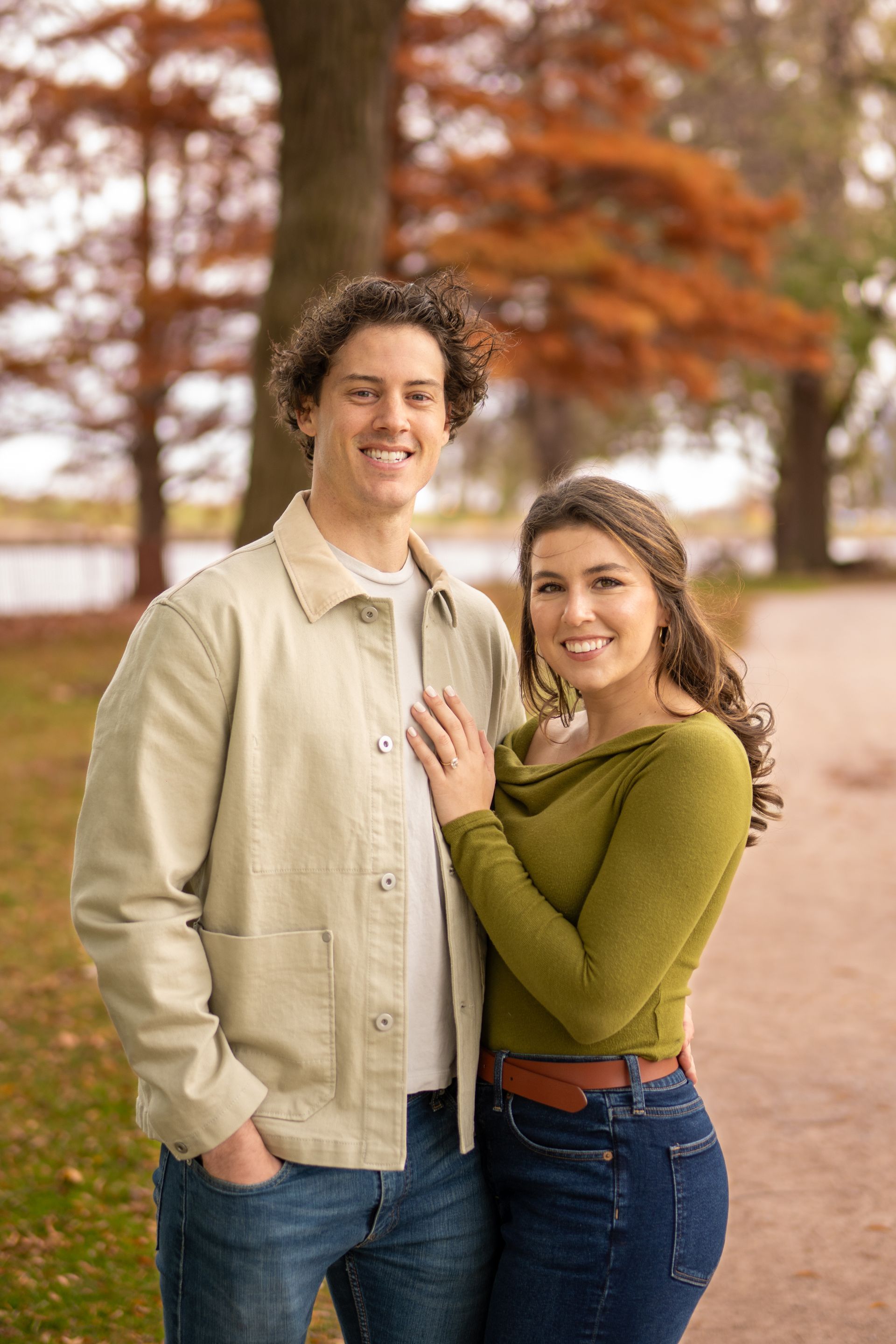 Couple poses outdoors; woman's hand on man's chest. Fall foliage in background.