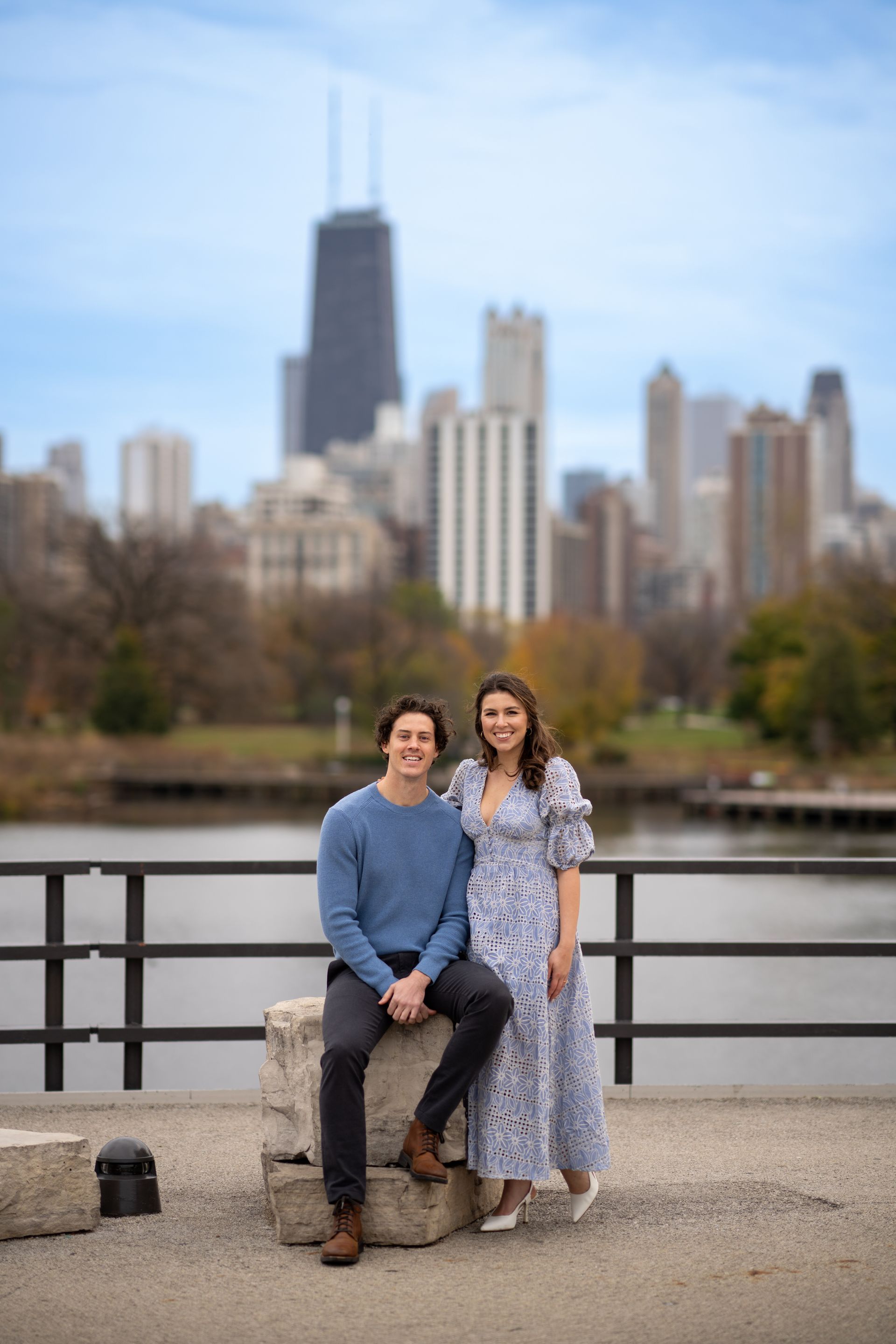 Couple poses by a Chicago skyline; the woman wears a blue floral dress, the man a blue sweater.
