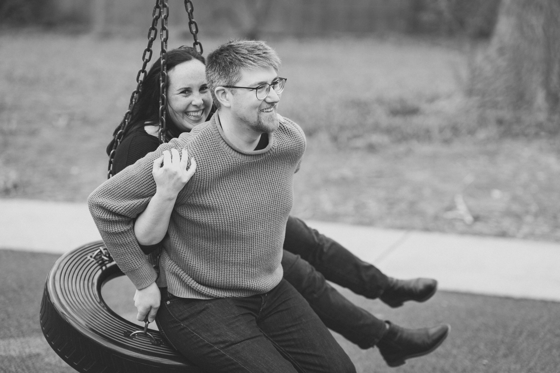 Couple on tire swing: Man swings with woman behind him. They smile in a park setting.