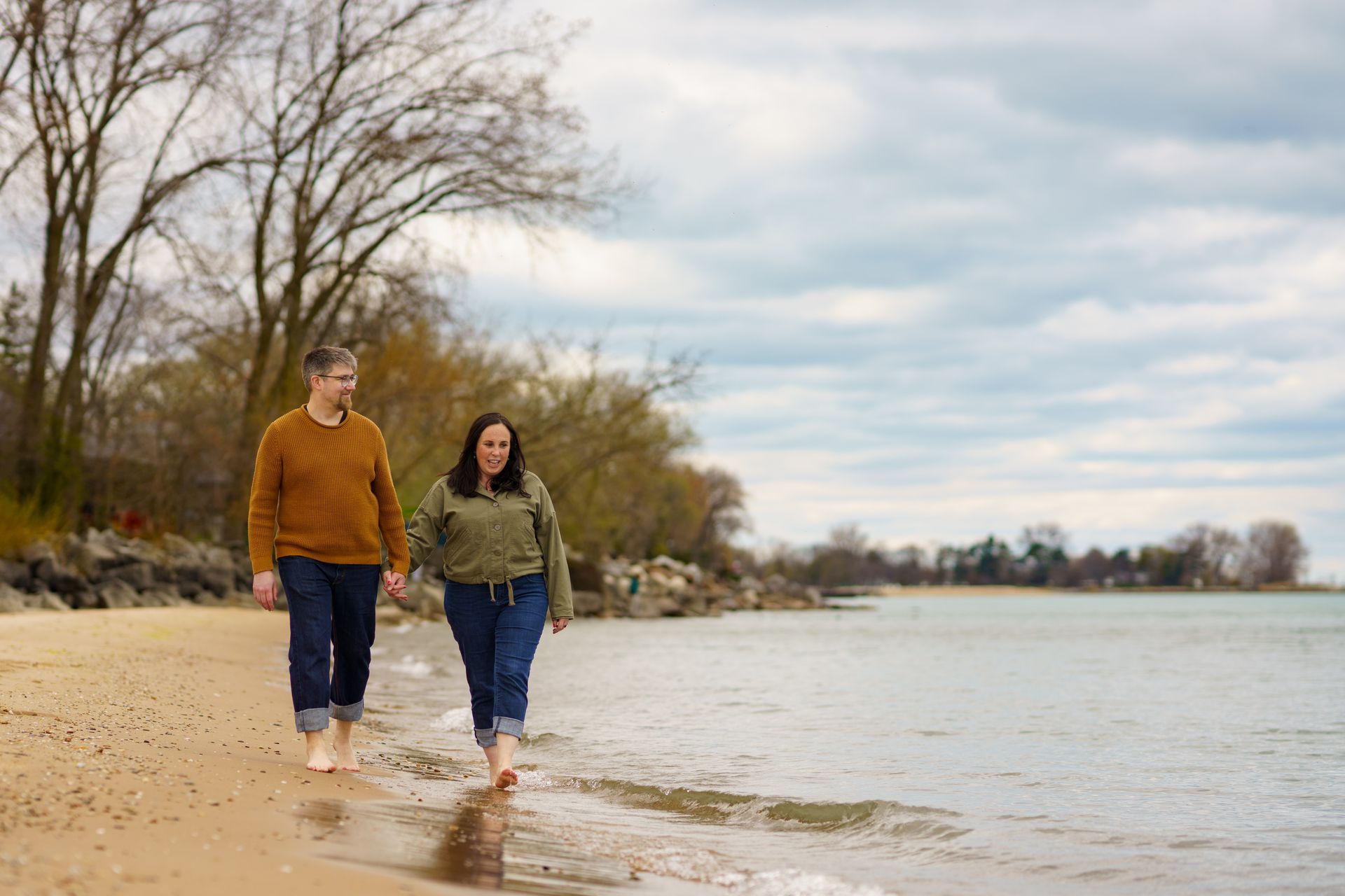 Couple walking barefoot along a sandy beach near water, cloudy sky overhead.