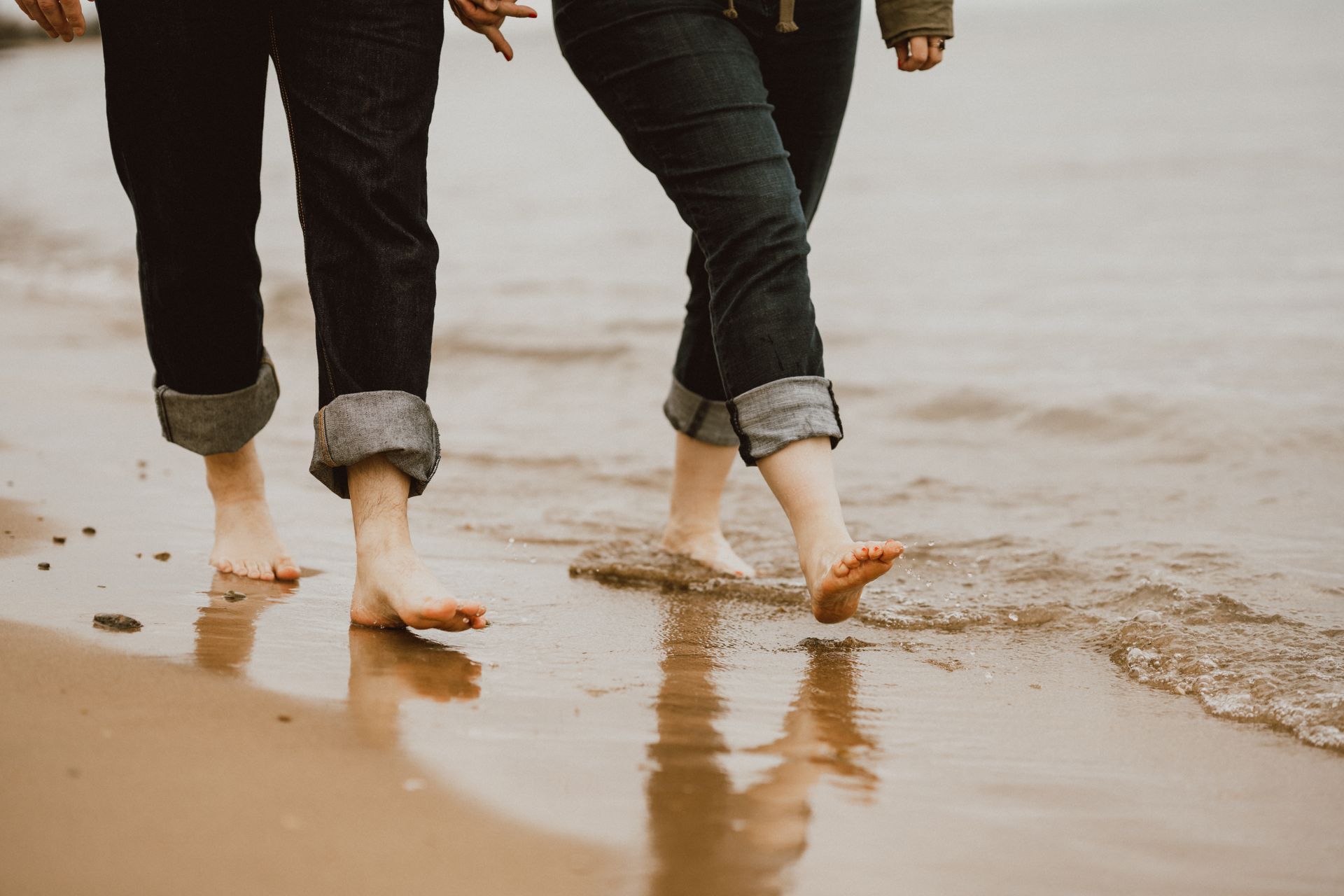 Three people walking barefoot on a beach, jeans rolled up, water at their feet.