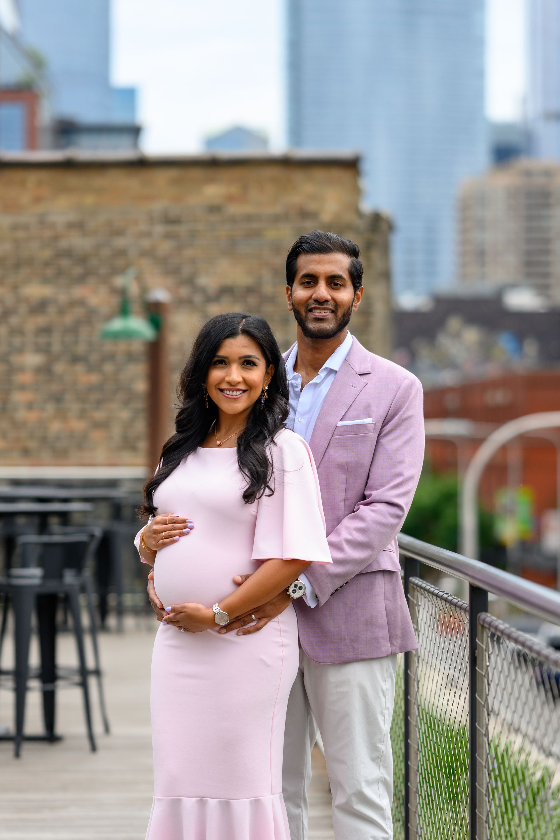 Pregnant woman in pink dress with man in a blazer, both smiling, outdoors on a patio with city buildings.