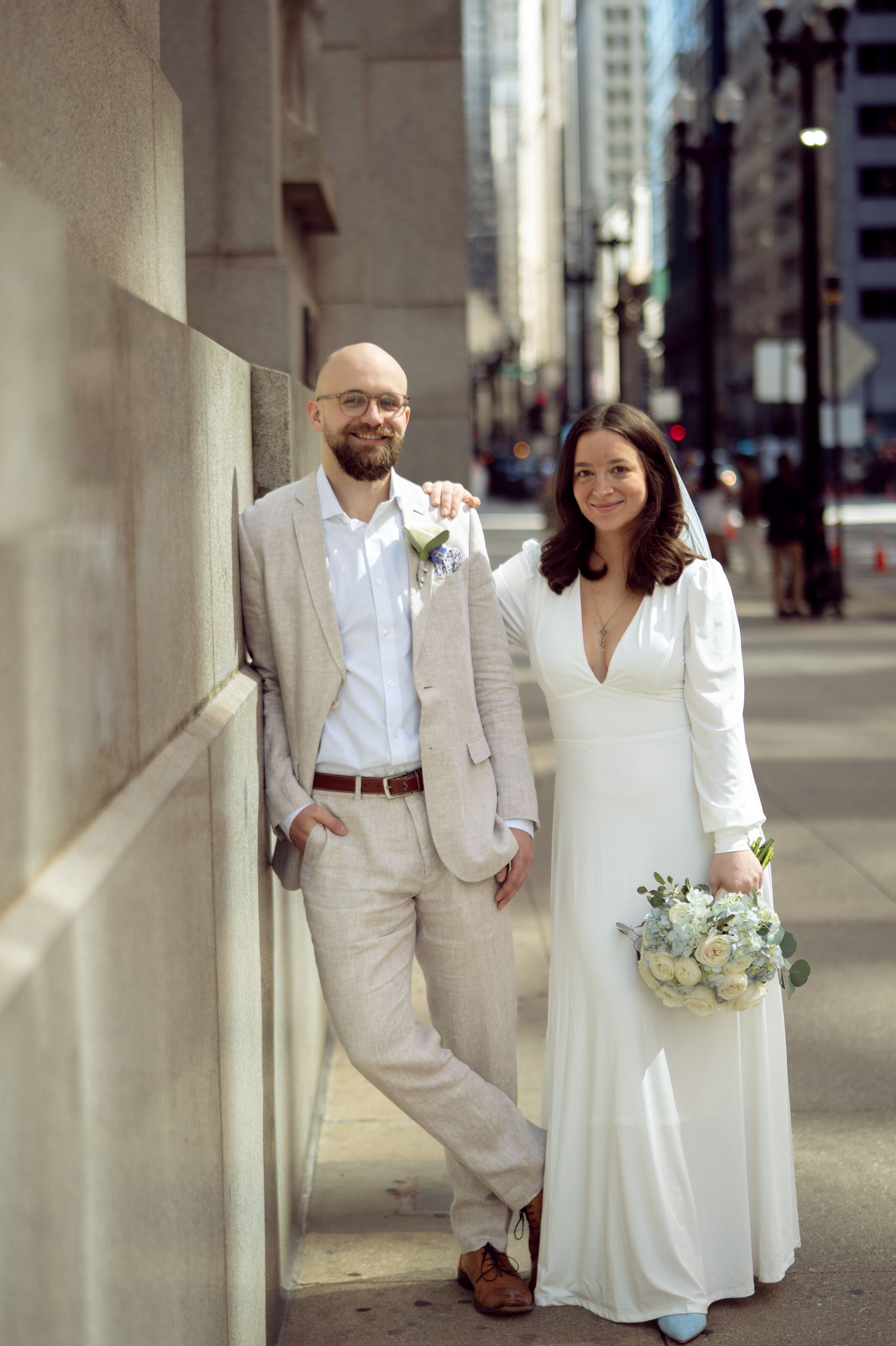Wedding couple posing outdoors by a stone wall; the woman wears a white dress and holds a bouquet.