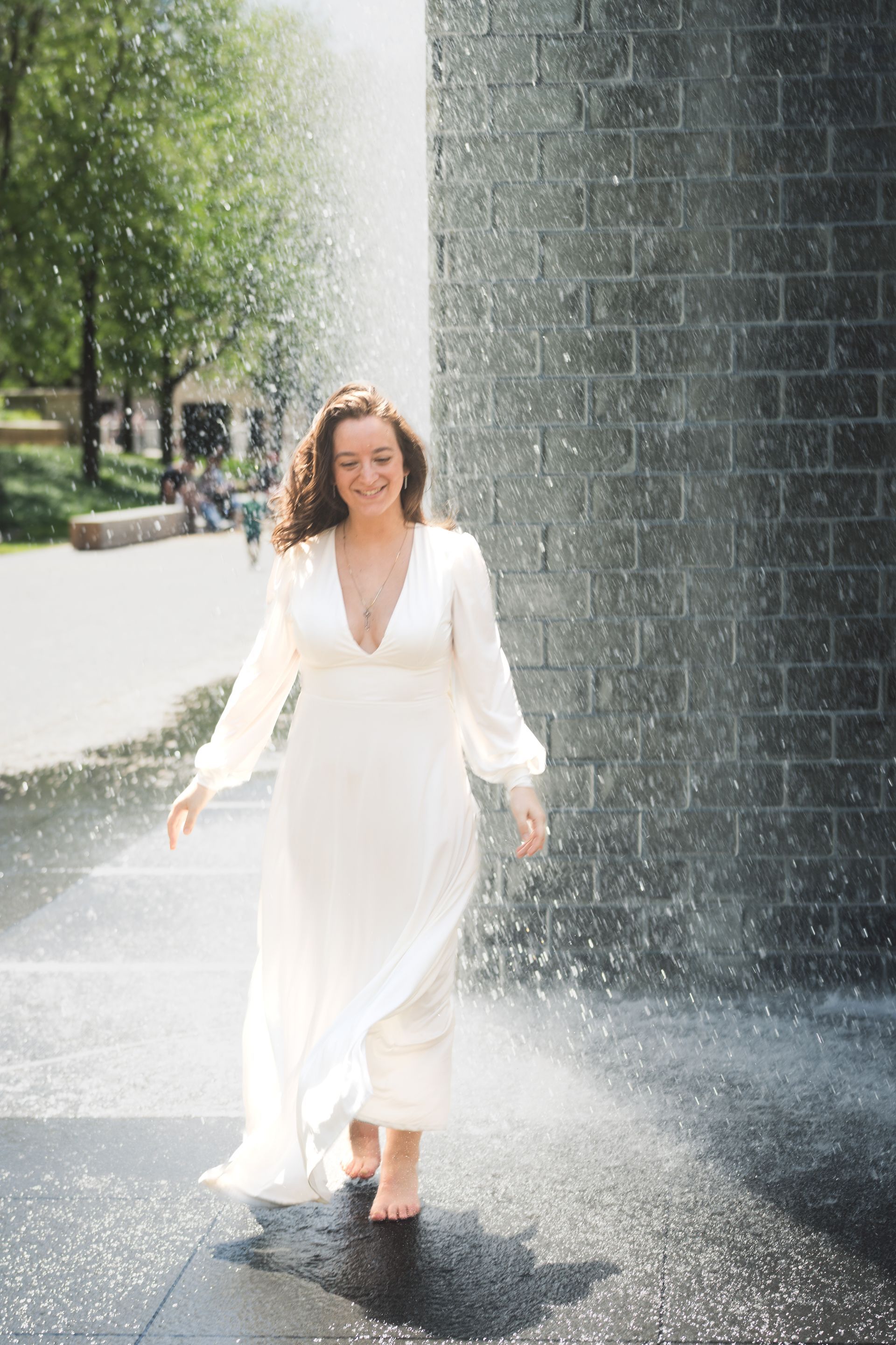 Woman in white dress, barefoot, joyfully walking through water fountain.