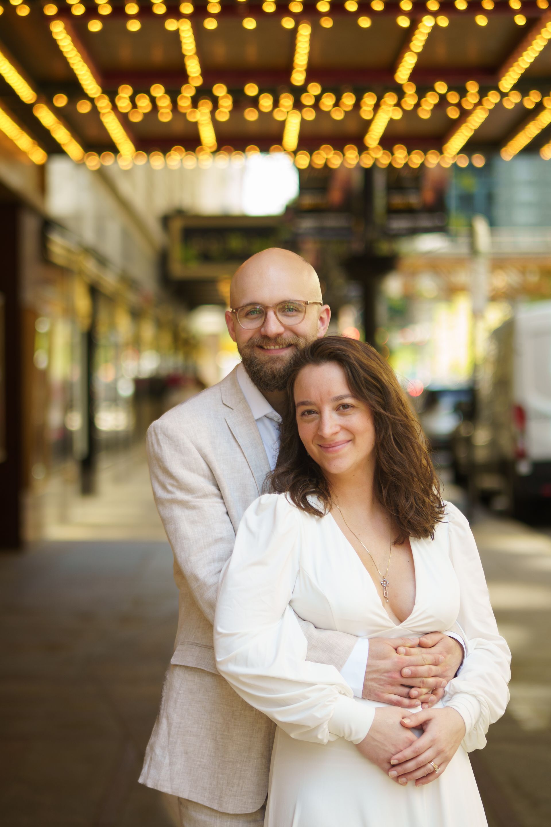 Couple embracing, smiling in front of a theater marquee. Woman in white dress, man in tan suit. Golden lights, city backdrop.