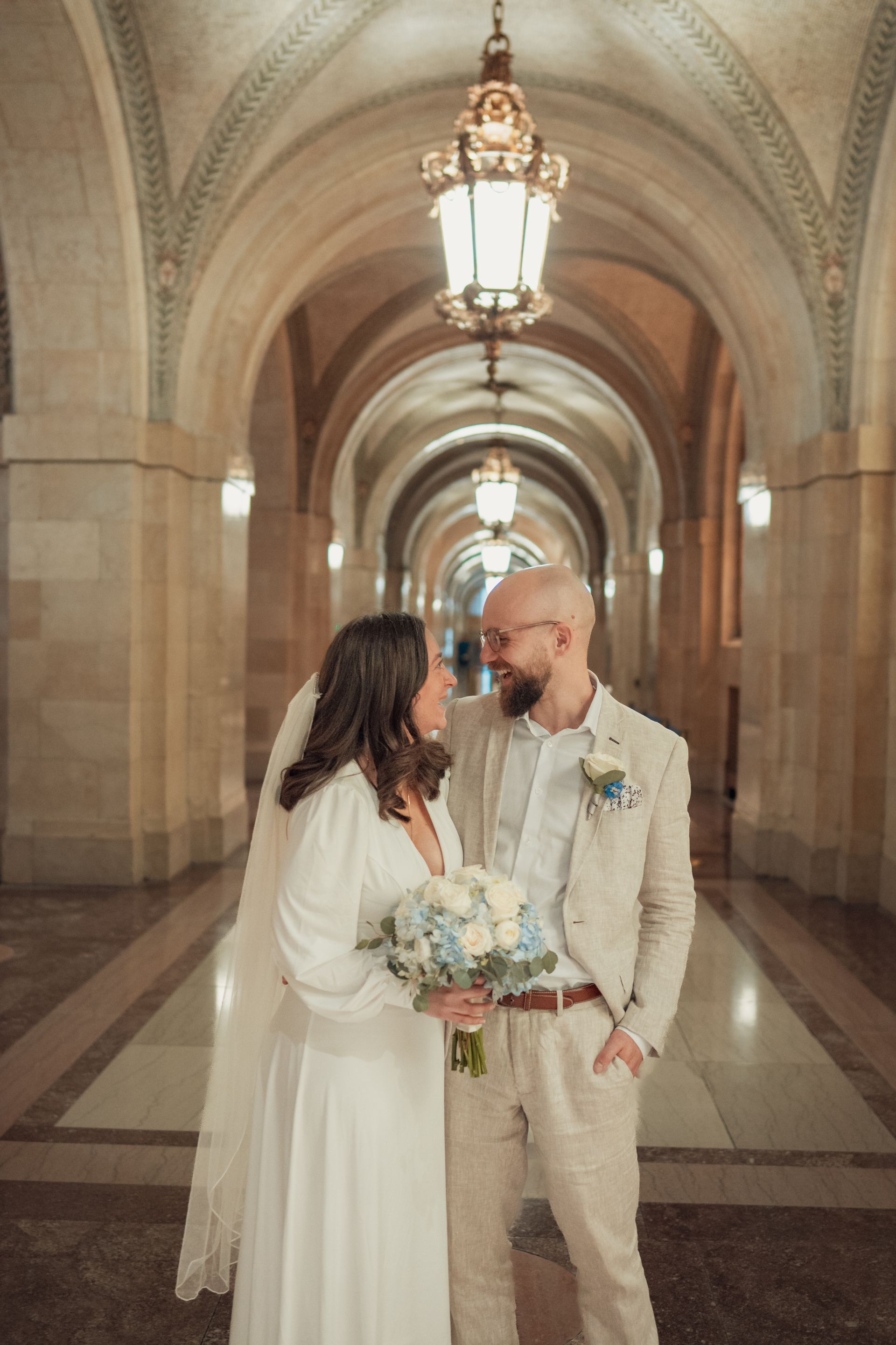 Couple in wedding attire gaze at each other in a stone arched hallway, lit by hanging lanterns.
