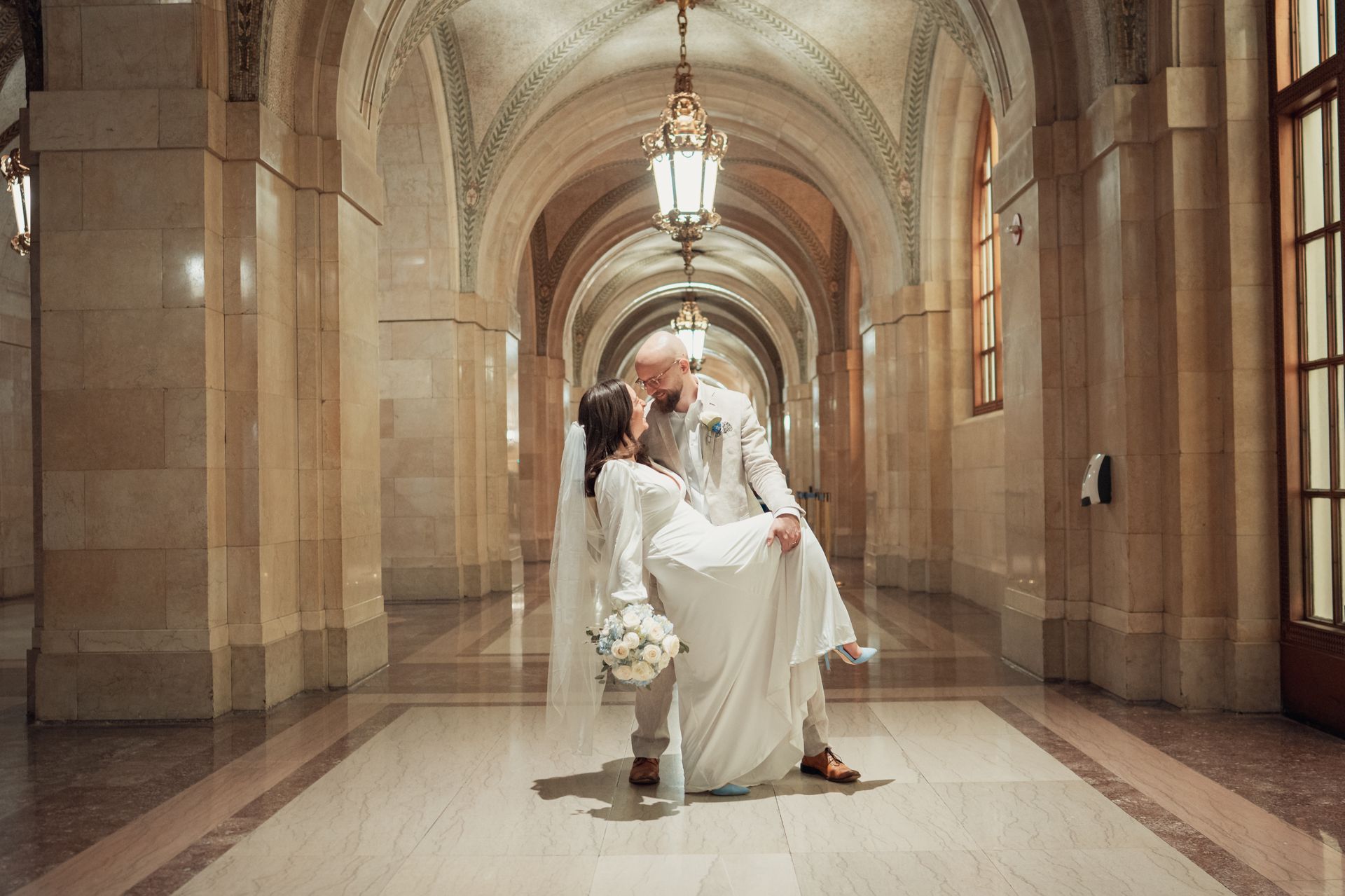 Groom holding bride in a white dress, hallway with arched ceiling, light fixtures, and stone walls.