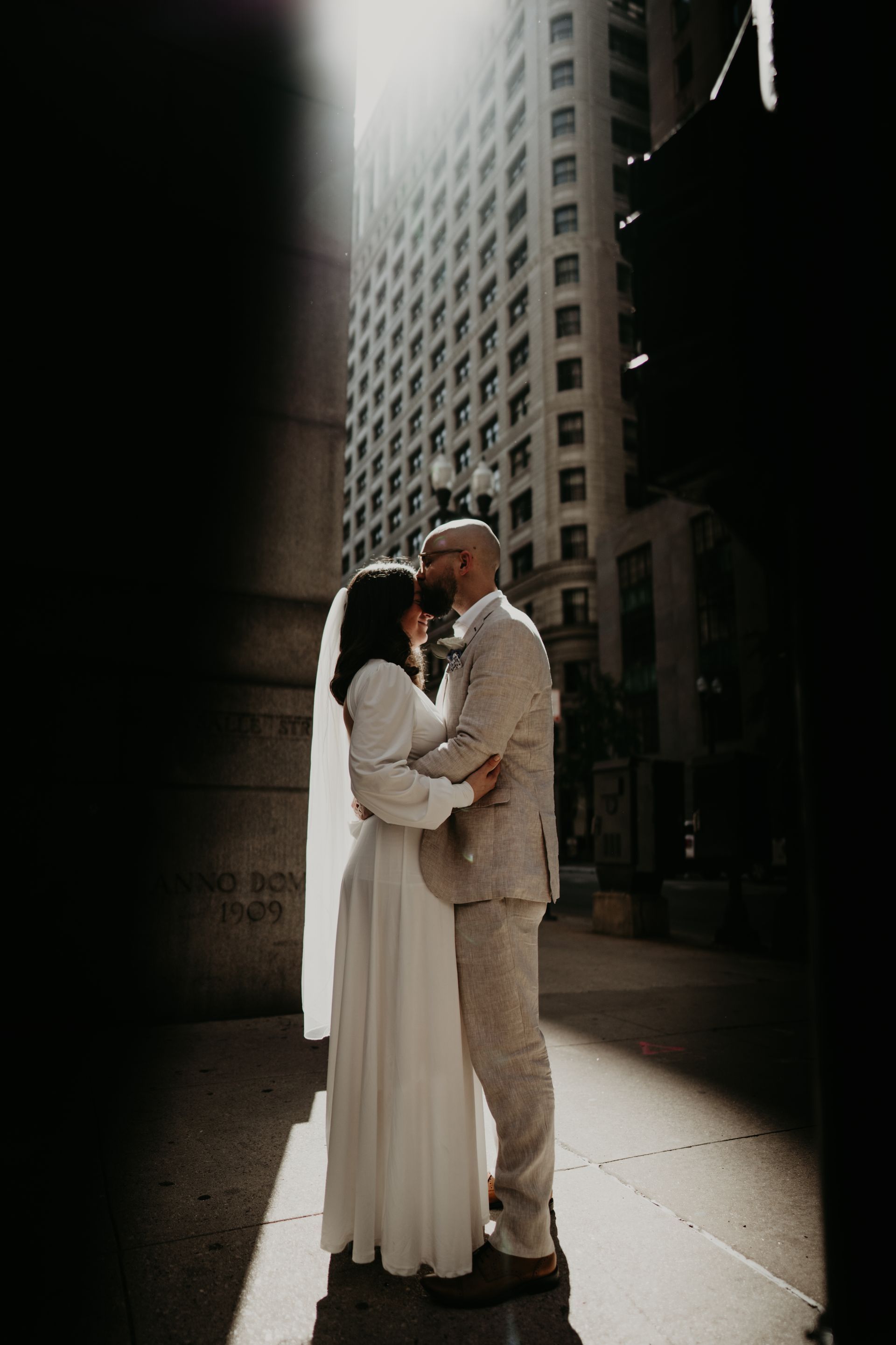 Couple embracing, groom kissing bride's forehead. Sunlit alley with tall building in background.