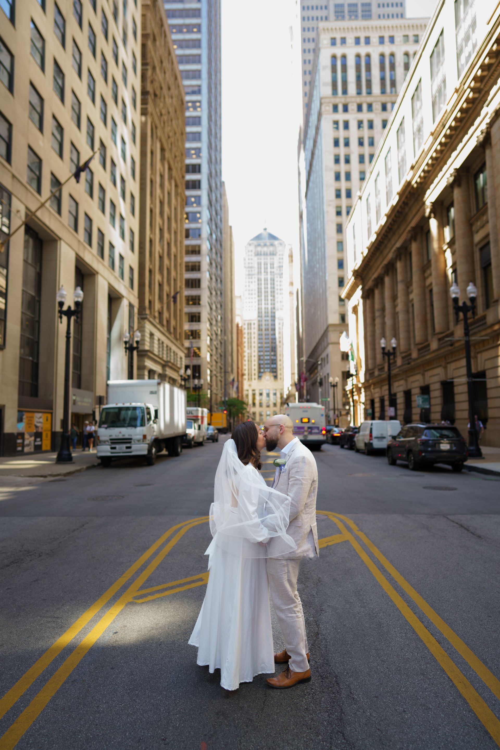 Couple kissing in a city street, center of the road, dressed in white outfits, tall buildings background.
