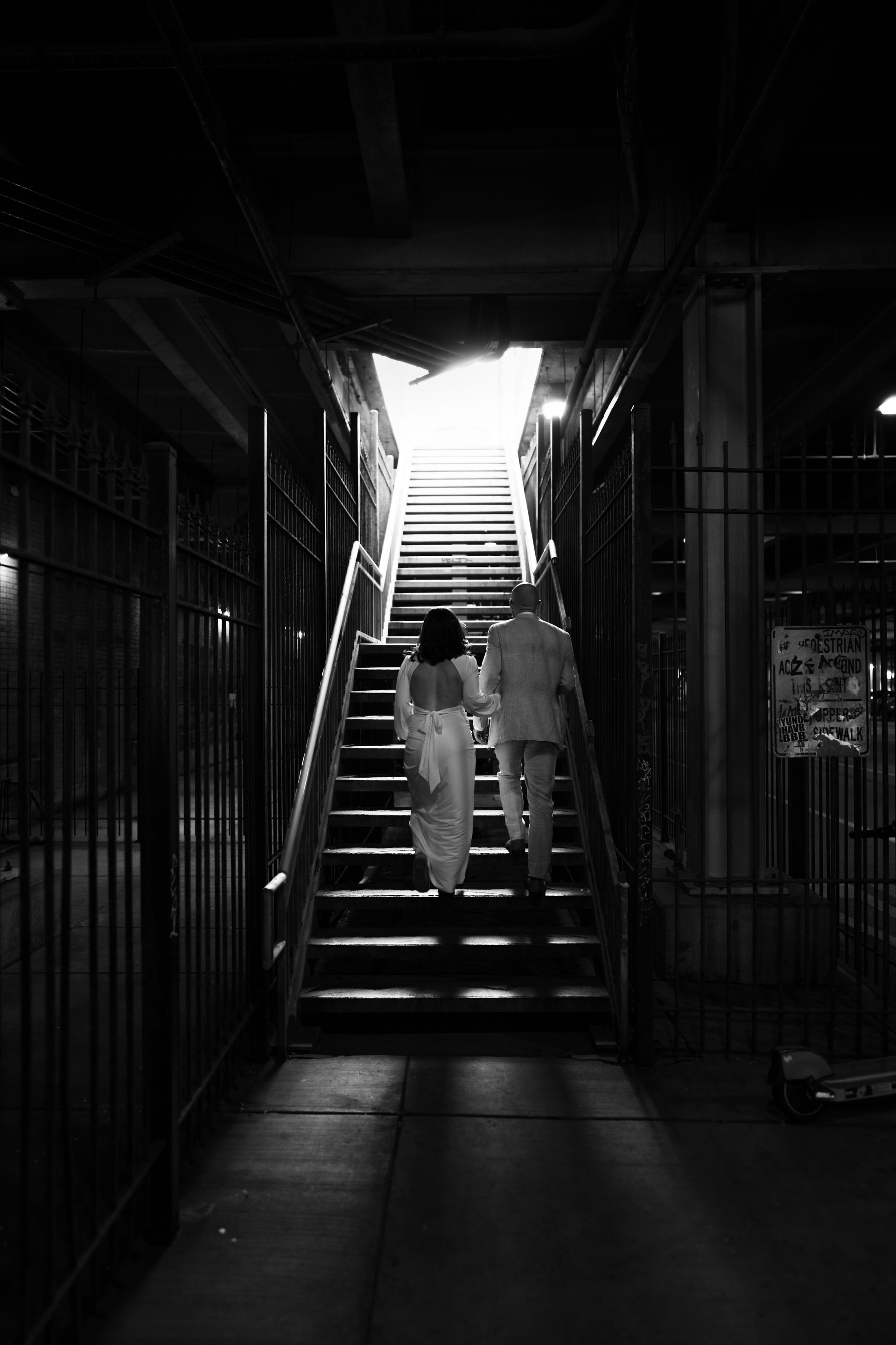 Couple ascending stairs towards bright light in a dark, caged passageway.