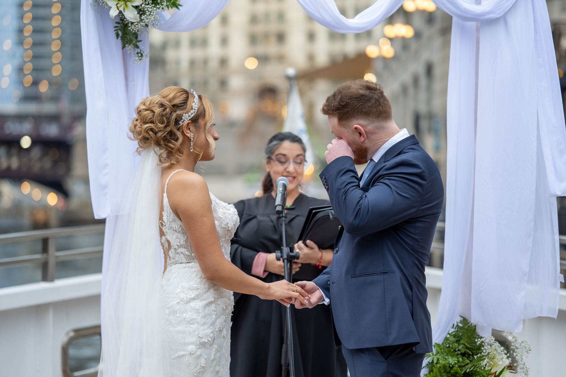Bride and groom at outdoor wedding ceremony; groom wiping his eye; white arch, river background.