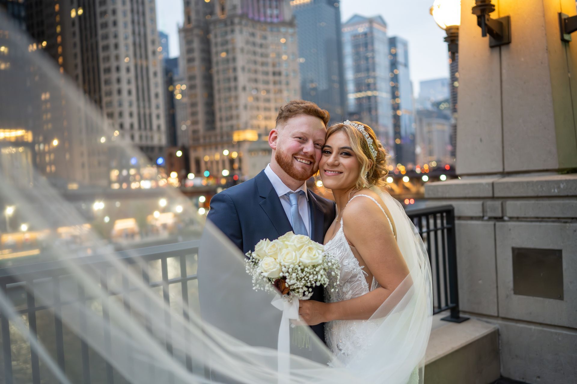 Couple on a bridge, smiling, with city skyline backdrop. Bride holds bouquet, veil blowing.
