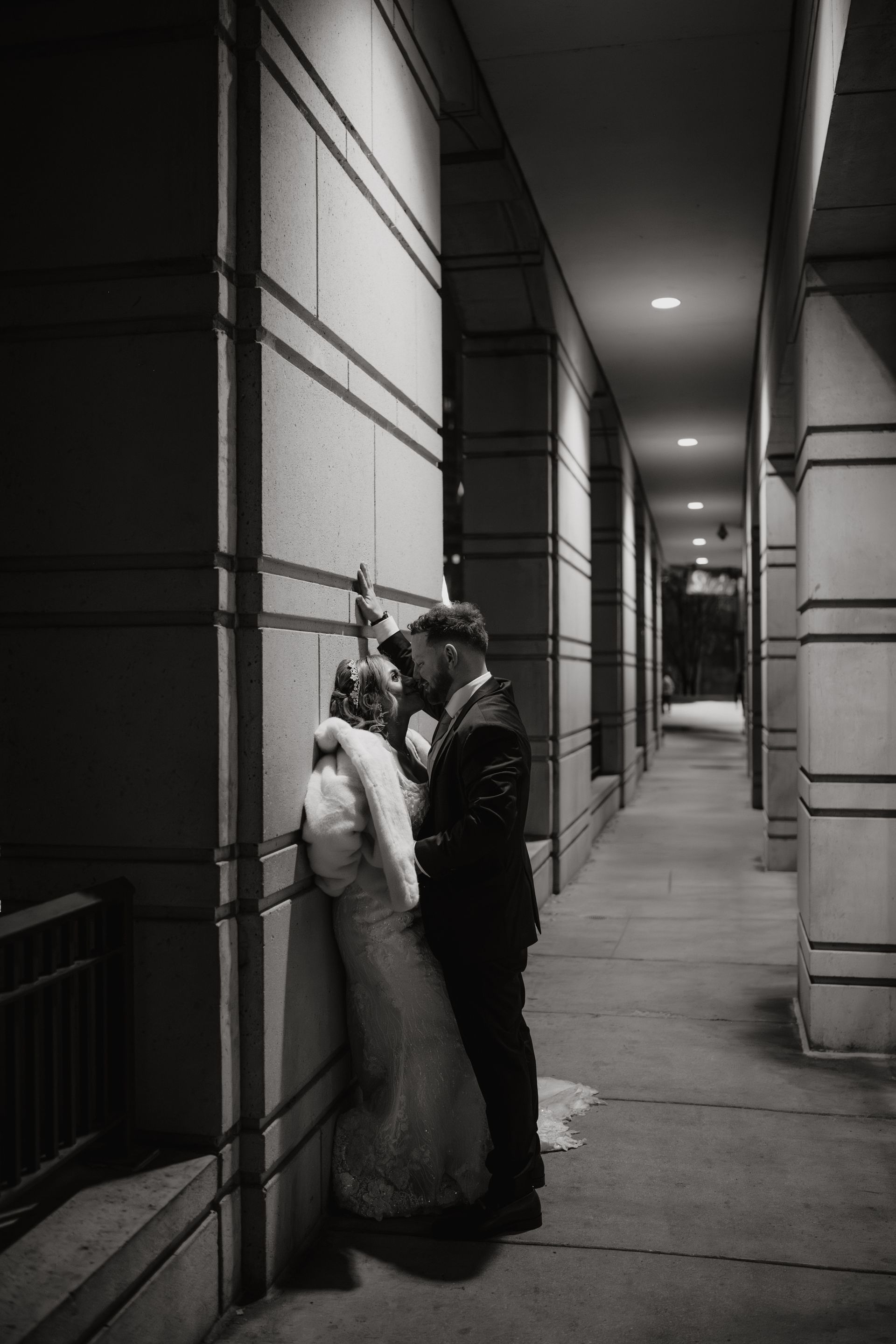 Newlyweds share a kiss against an outdoor building wall, illuminated by overhead lights.