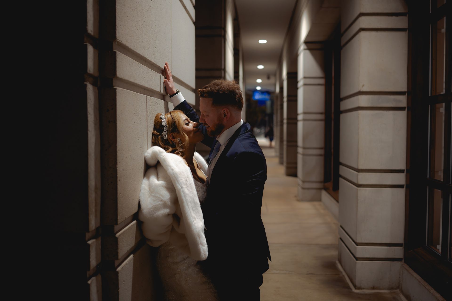 Couple kissing, bride in fur wrap leans against wall, groom in suit. Dark hallway, overhead lights.