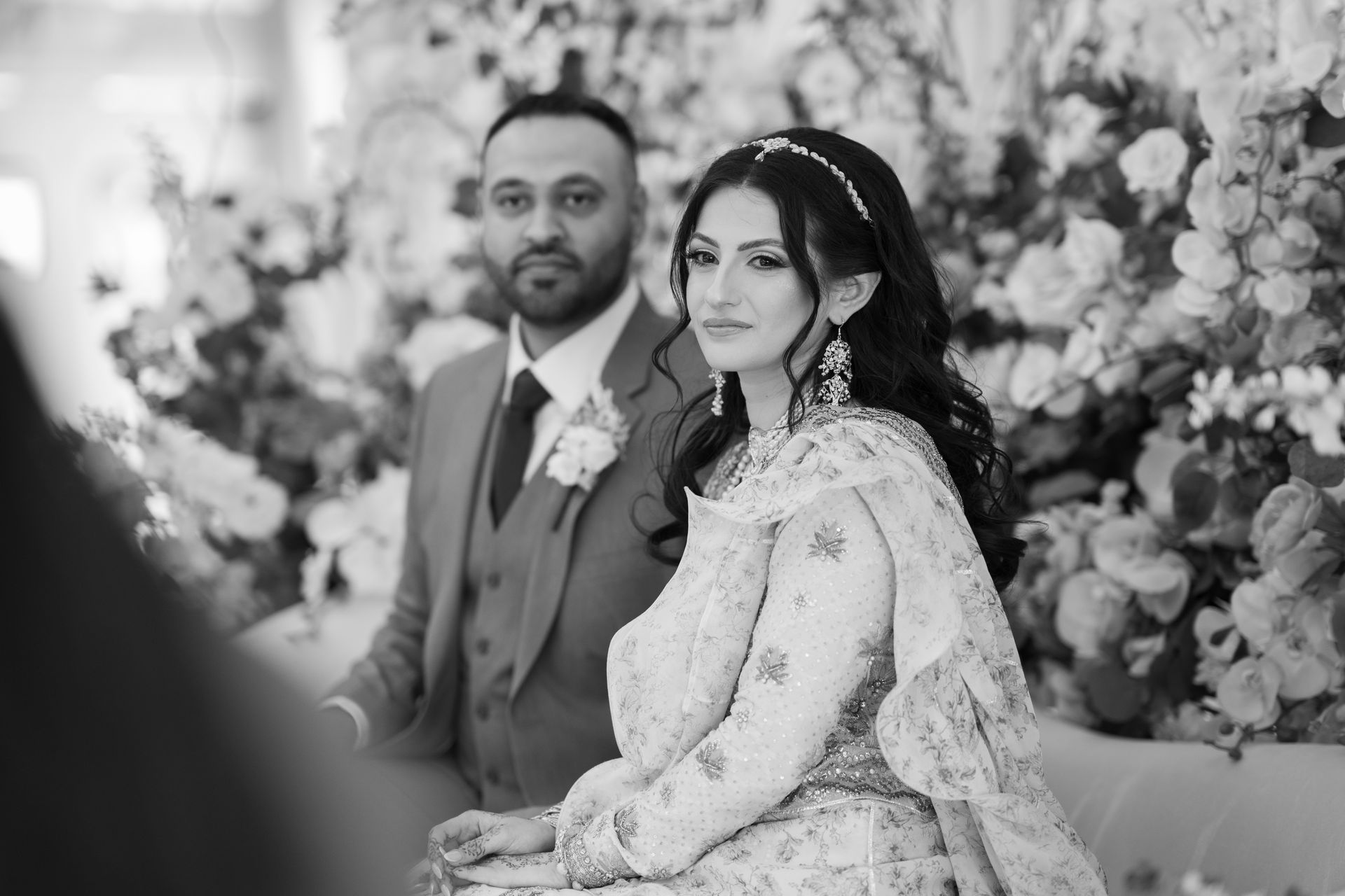 A couple in formal attire sit indoors amidst floral decorations; the woman wears a headpiece and ornate garment, while the man wears a suit.