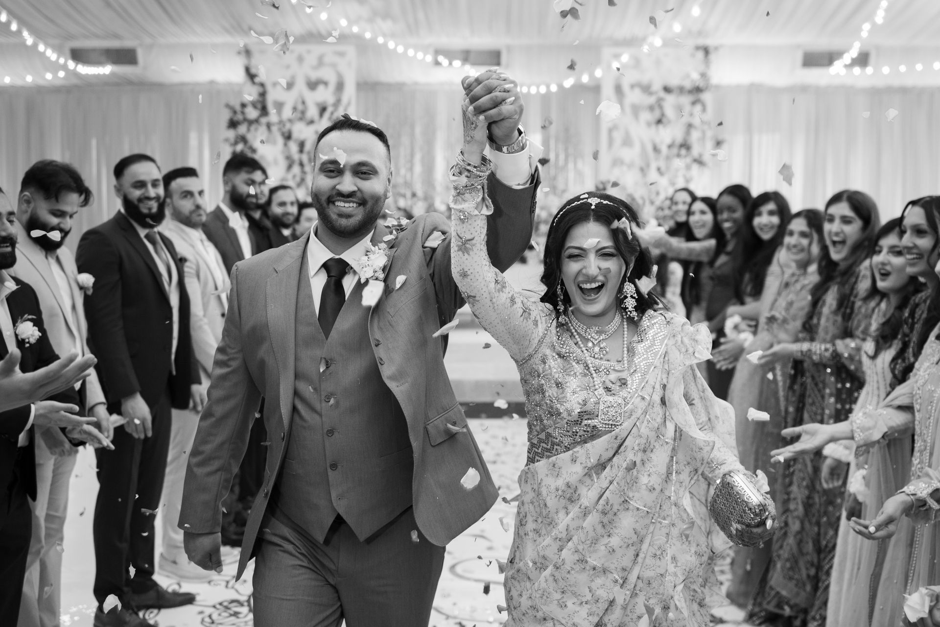 Newlyweds walk hand-in-hand, smiling, amid confetti and cheering guests at a wedding reception.