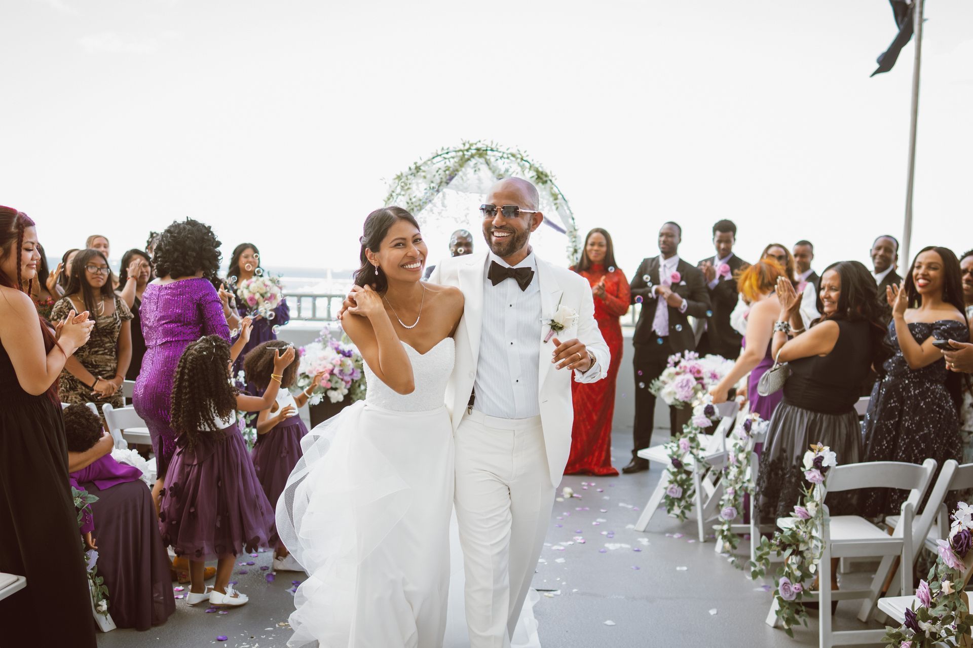 Newly married couple walking down the aisle, surrounded by guests clapping. Wedding on deck, floral arch.