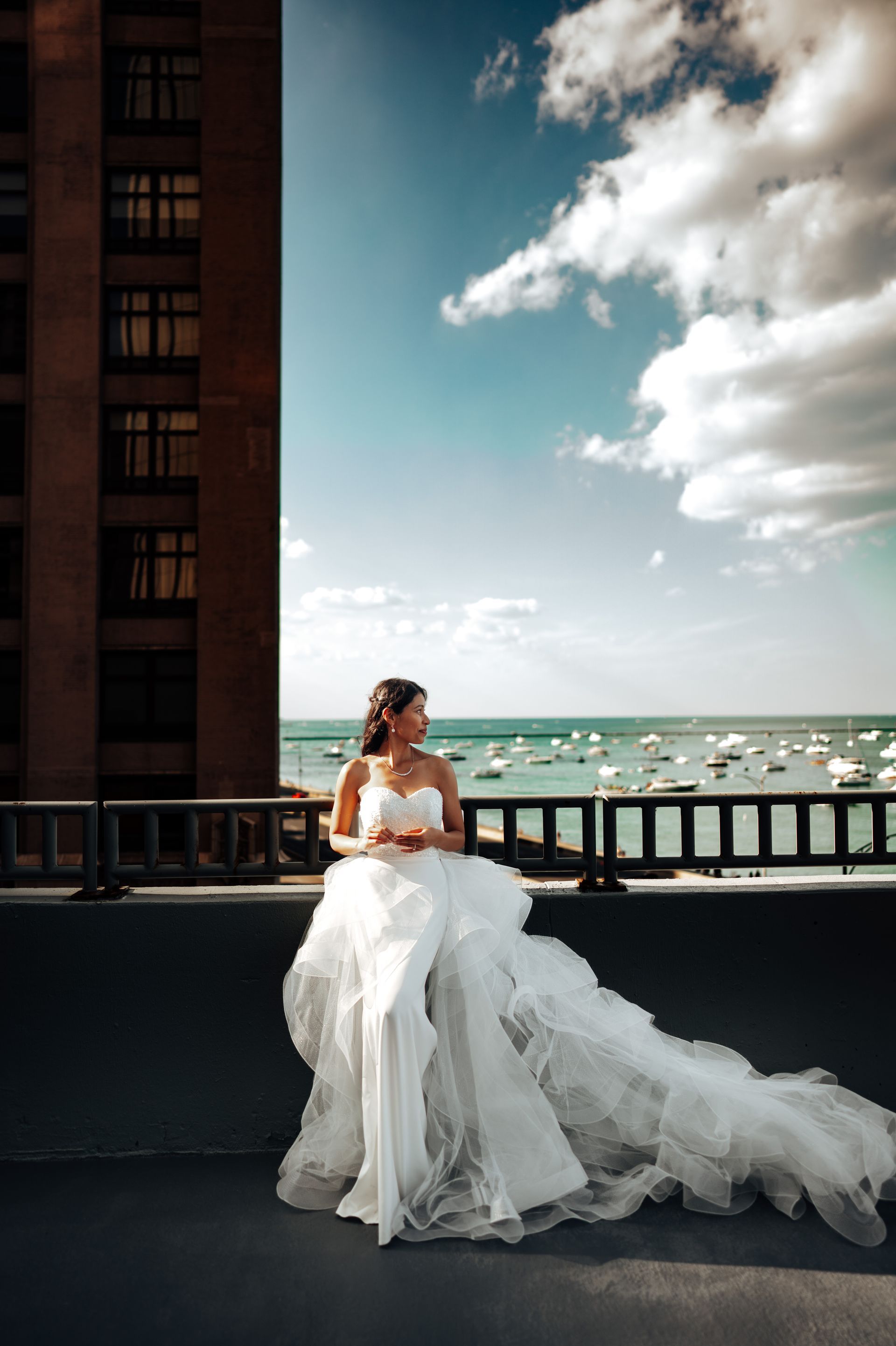 Bride in flowing white gown on a rooftop with ocean view and boats, blue sky with clouds.