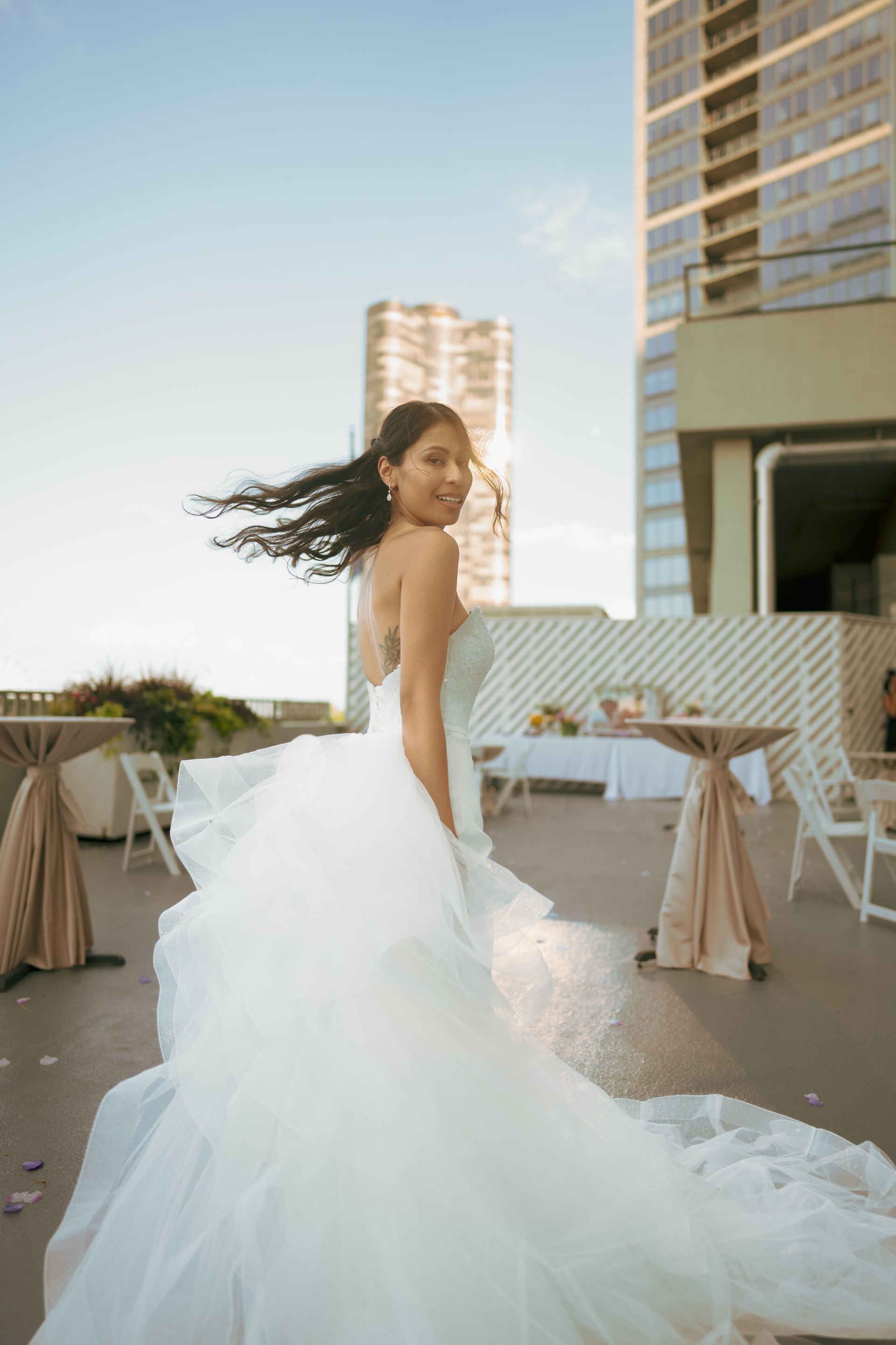 Bride in white gown twirls on rooftop, looking back over shoulder. Sunlit background, city view.