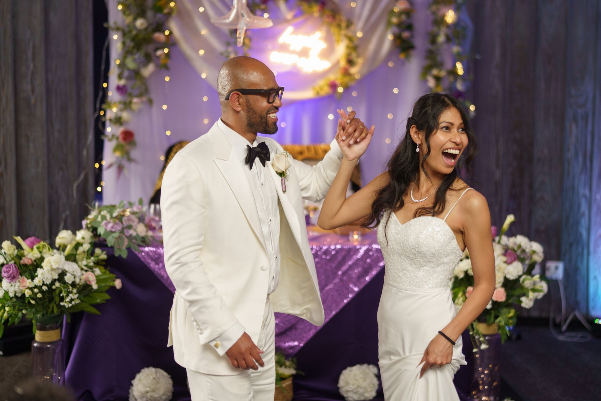 A newlywed couple celebrates at their wedding reception. The bride wears a white gown, and the groom a white tuxedo.