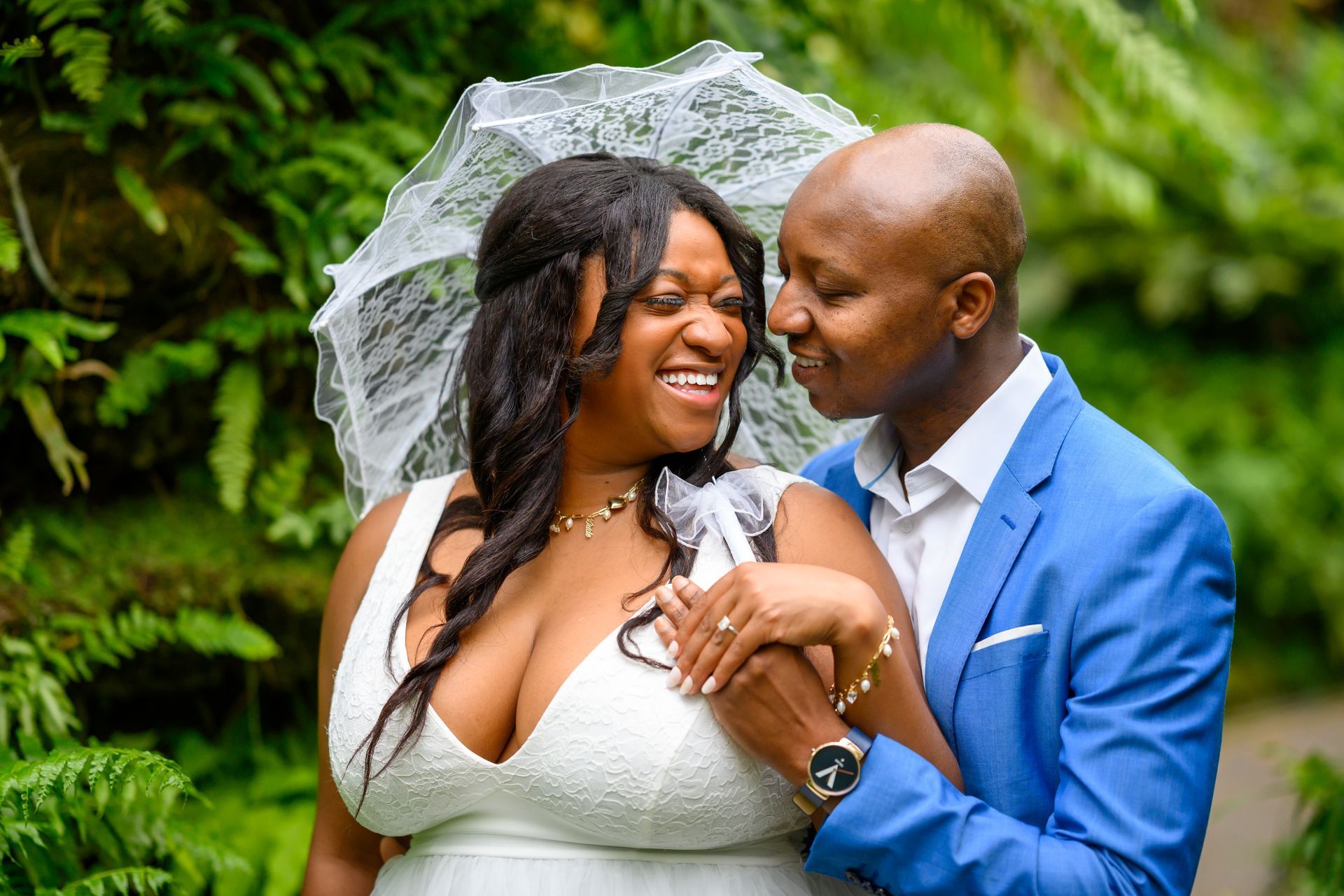 Bride and groom embrace, smiling. Bride in white dress and parasol. Groom in blue suit. Green foliage backdrop.