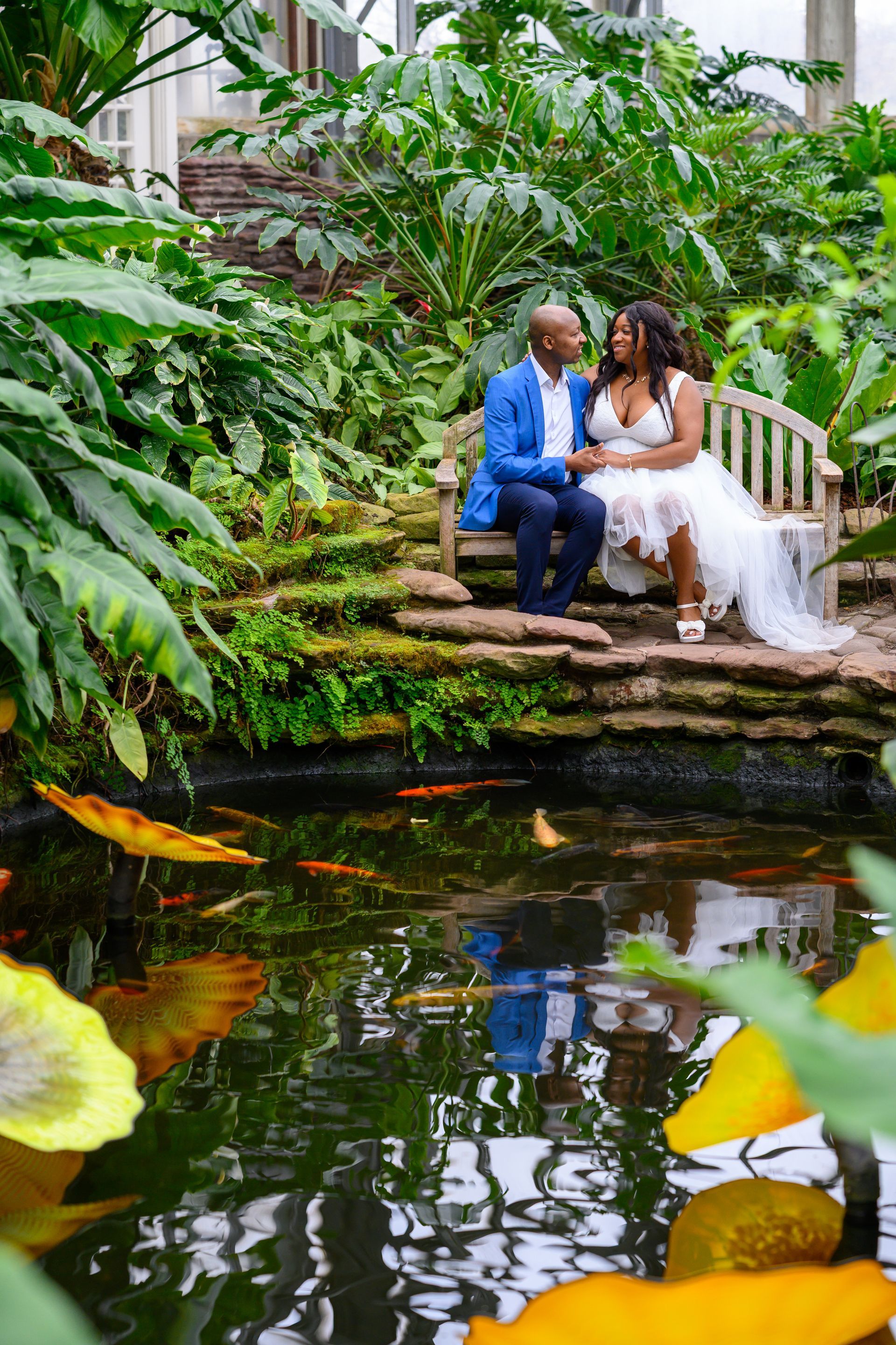 Couple seated on a bench, looking at each other, beside a koi pond, surrounded by lush greenery.