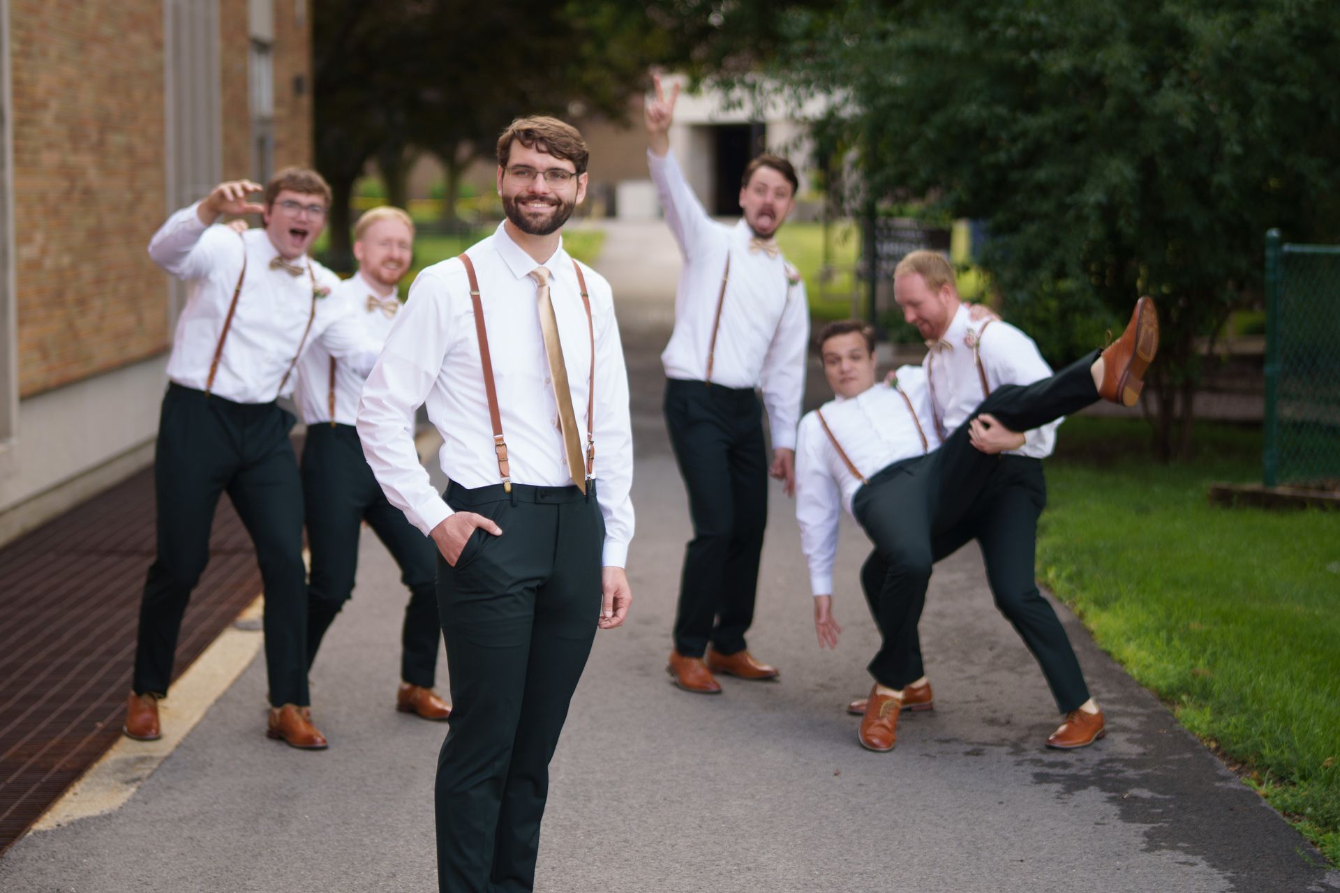 Groom and groomsmen posing on a path. They wear white shirts, suspenders, and dark pants, with varied, playful expressions.
