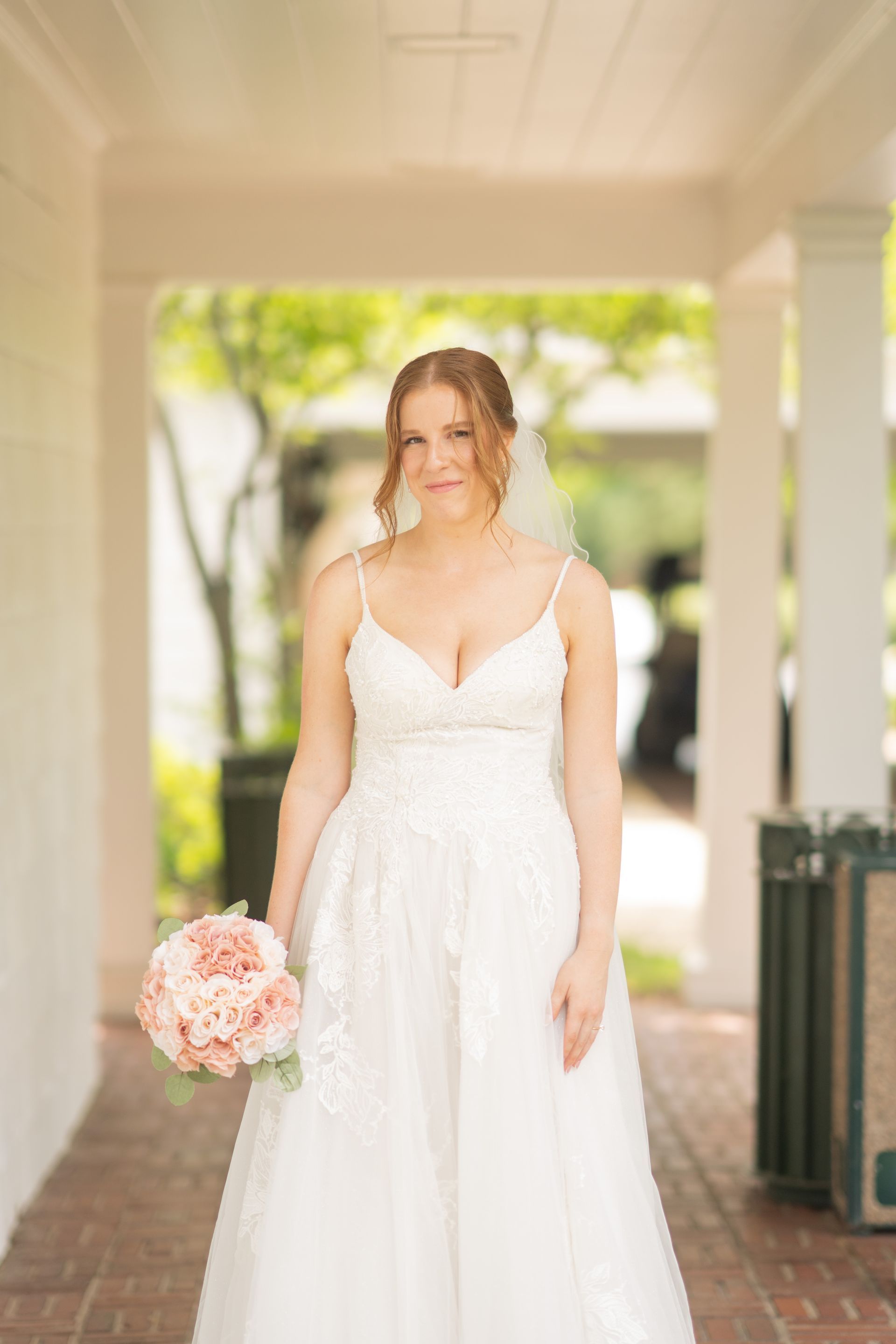 Bride in white dress, holding pink bouquet, standing in a brick corridor with greenery in the background.