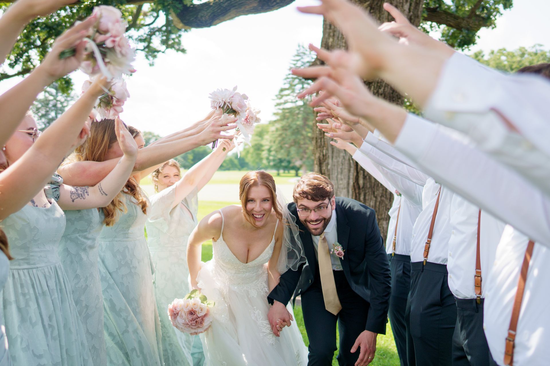 Bride and groom run through a raised-arm tunnel created by wedding party. Outdoors, in a sunny setting.