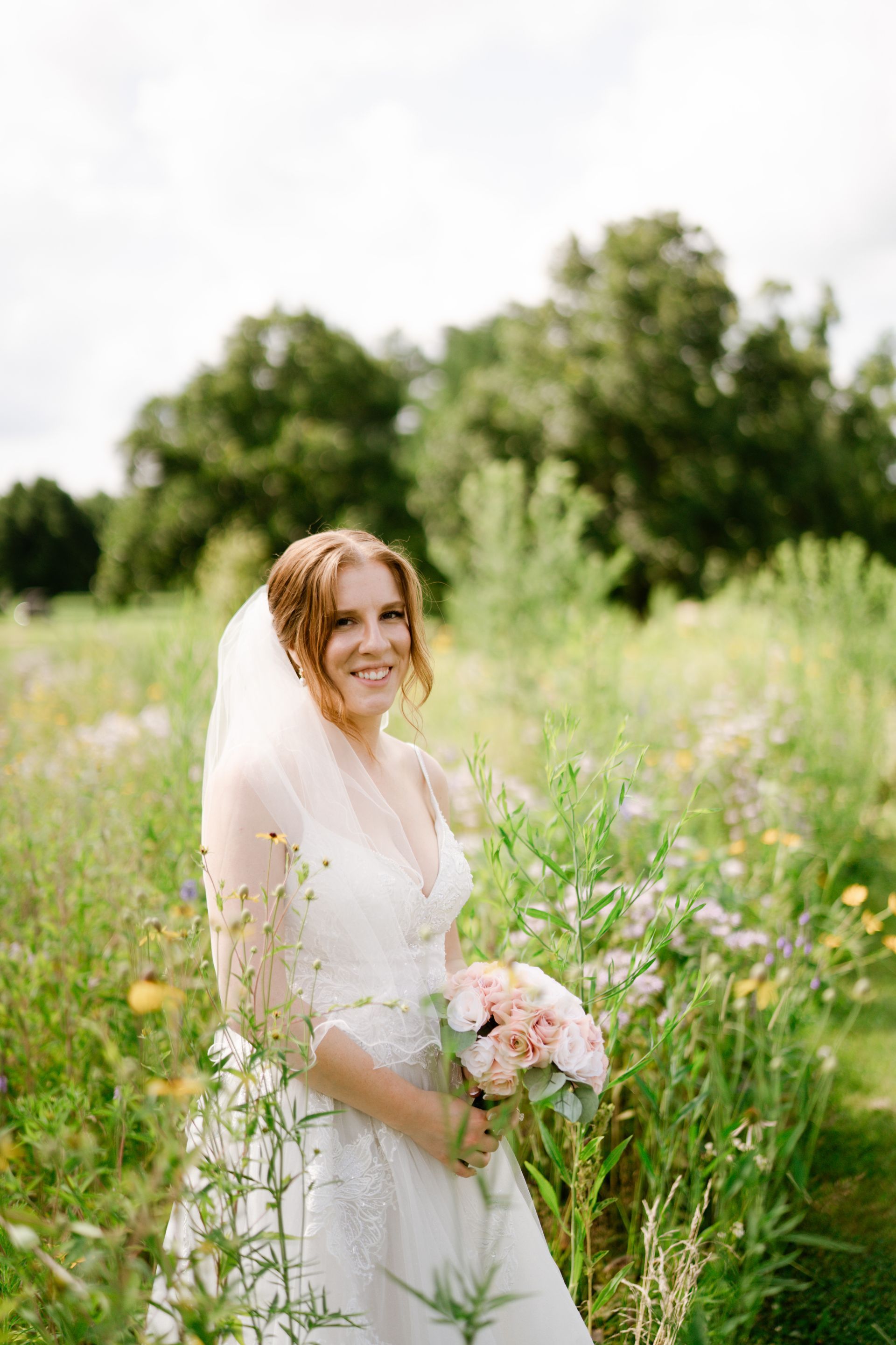 Bride in a white dress and veil holds a bouquet in a wildflower field; smiling.